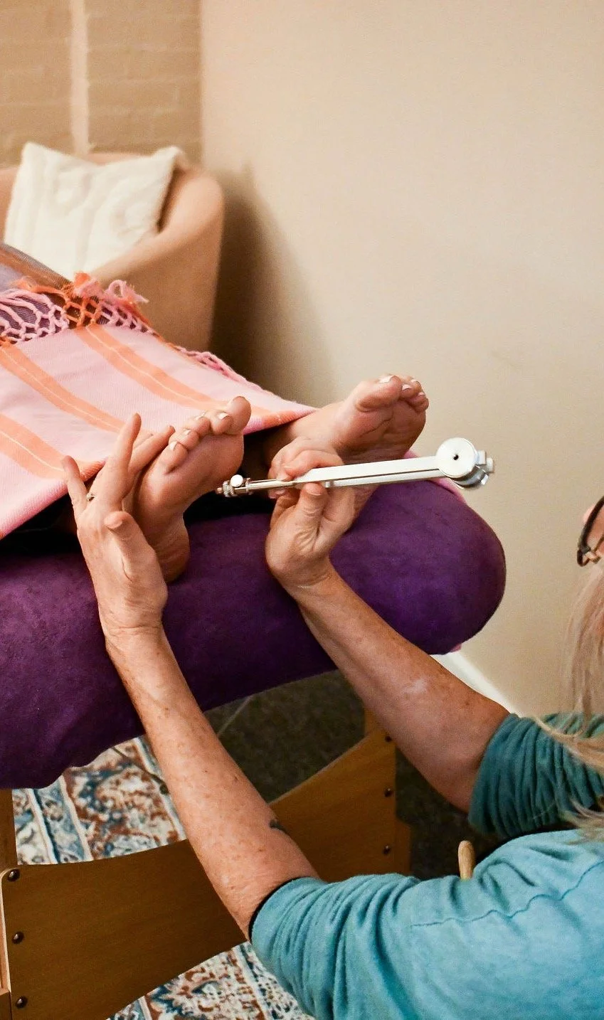 A person’s crossed arms legs raised in front of a colourful, abstract circular background with radiating lines, resembling a psychedelic or cosmic design, with three purple tuning forks next to her.