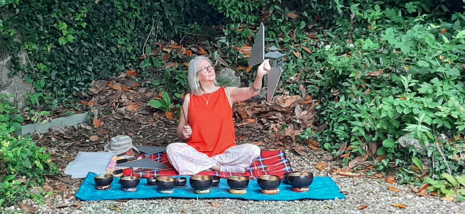 A woman sitting cross-legged on a blanket outdoors, playing a sound wave, with singing bowls and potted plants around her, surrounded by green foliage.
