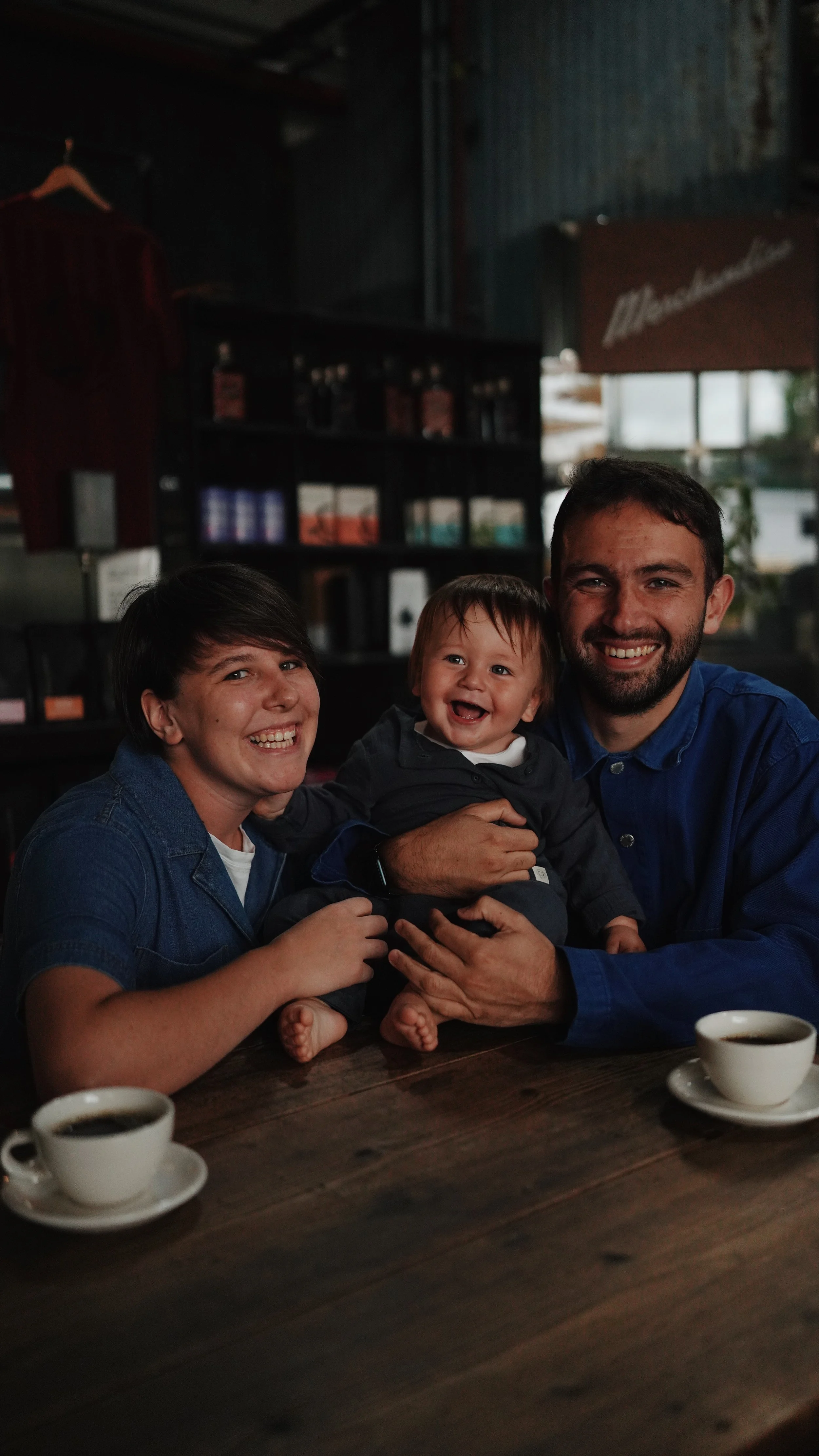 A smiling family of three, including a woman, a man, and a young child, sitting at a table in a cozy cafe with coffee cups in front of them. The woman and man are holding the child, who is laughing.