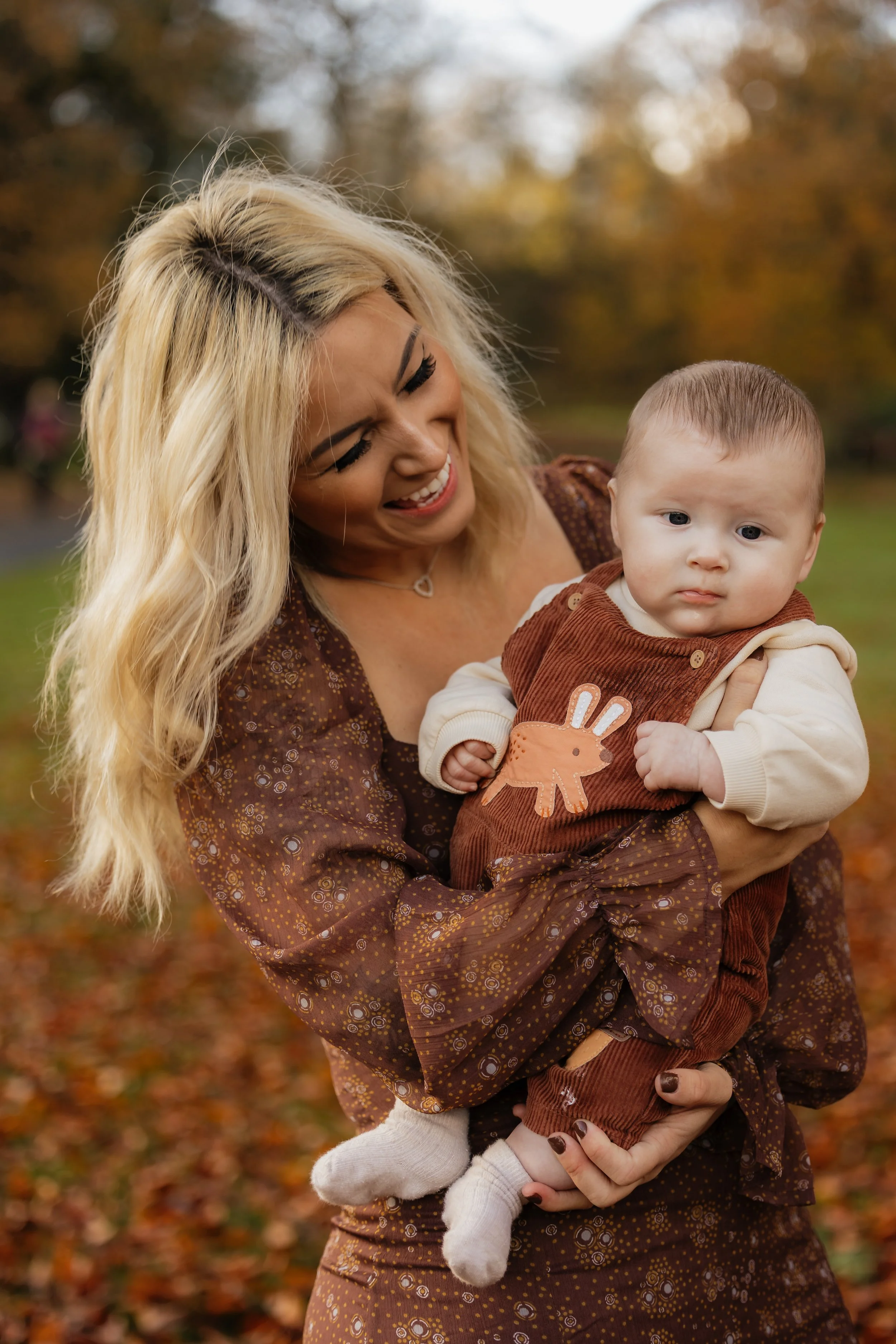 A woman holding a baby outdoors in a park during autumn with fall leaves on the ground and trees in the background.