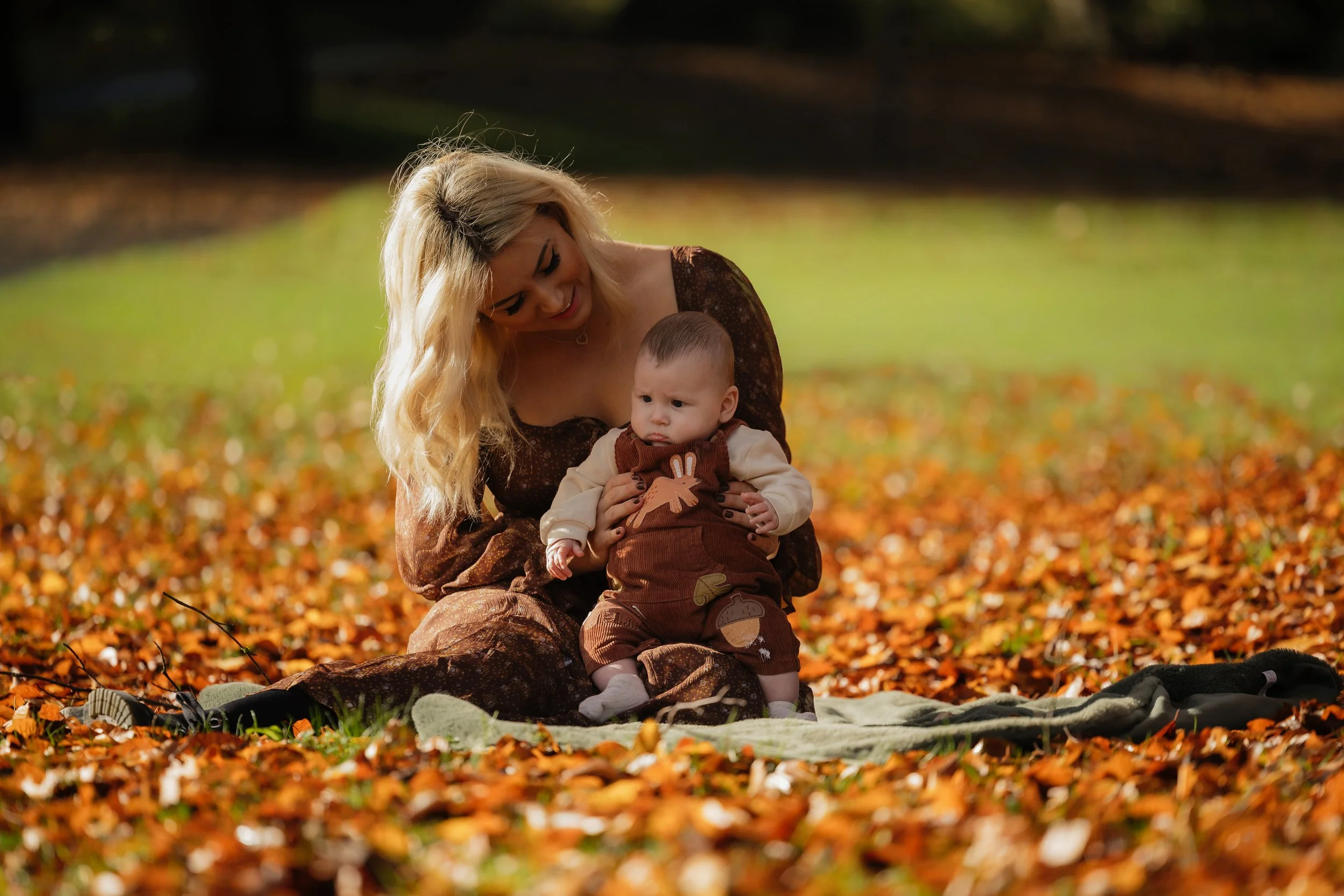 A woman and a baby sitting outdoors on a blanket among fallen autumn leaves, with trees in the background.