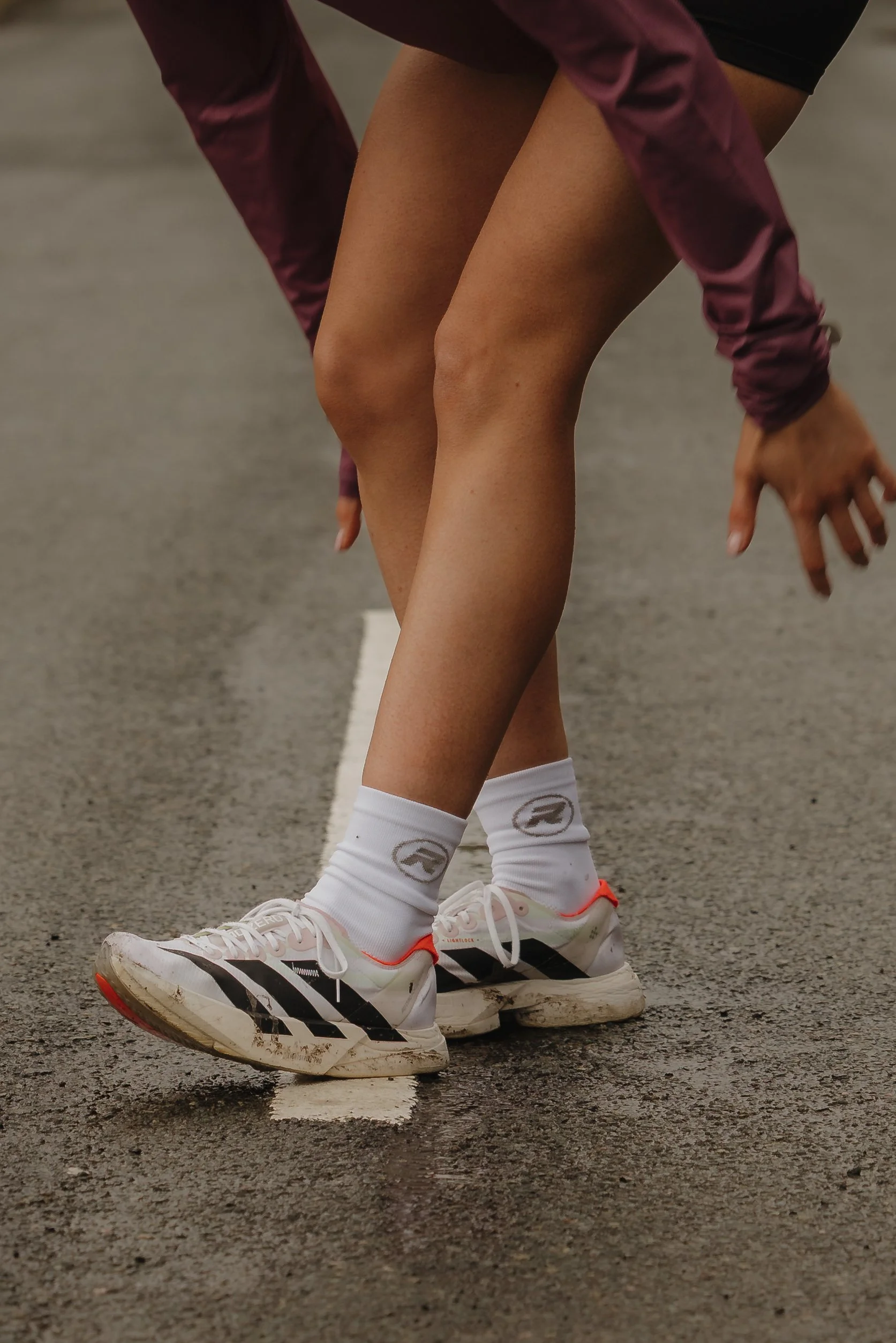 A person wearing running shoes and white socks crouches on the road at the start of a race, their hands touching the ground.