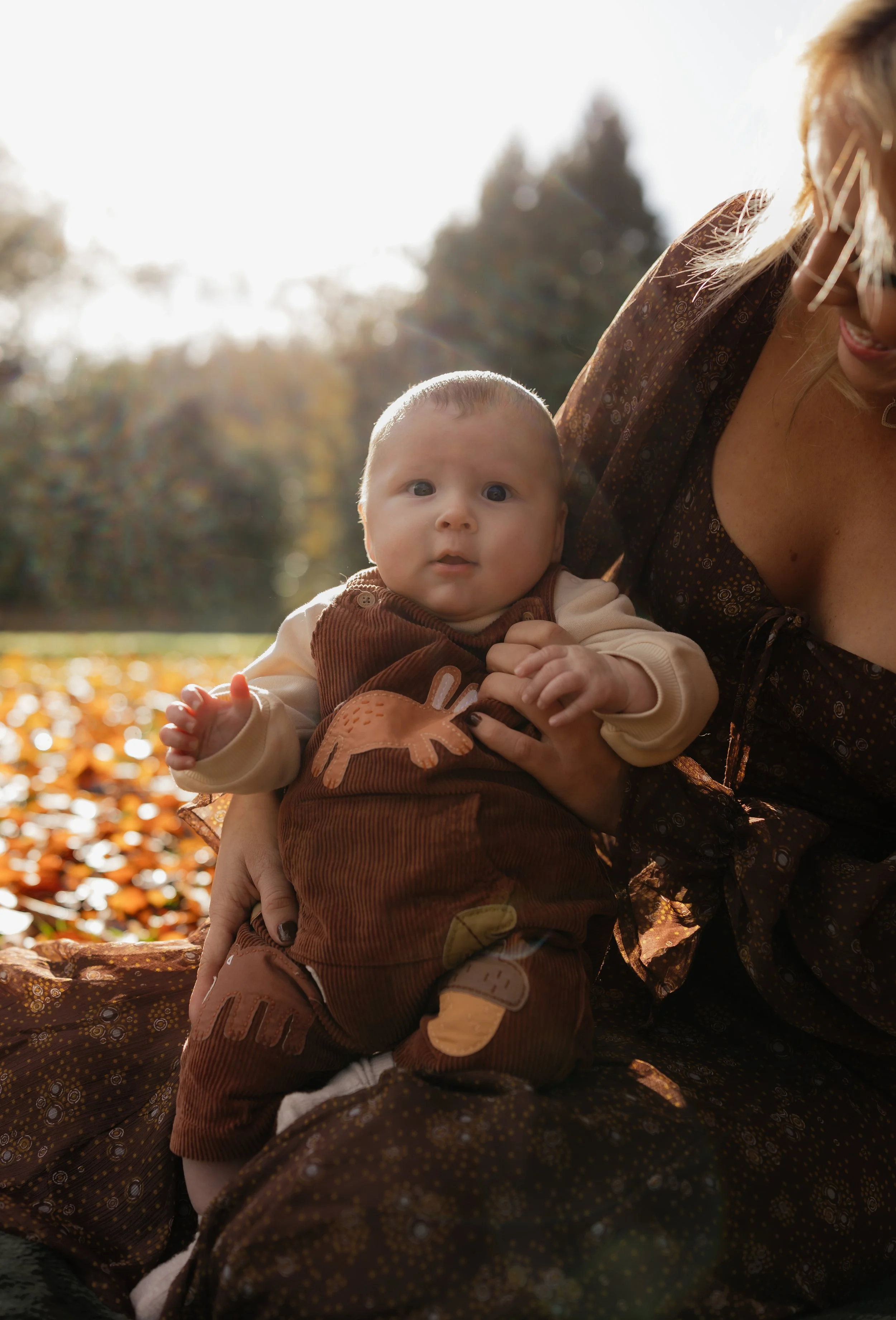 A woman holding a baby outdoors in an autumn park with fallen leaves and trees in the background.