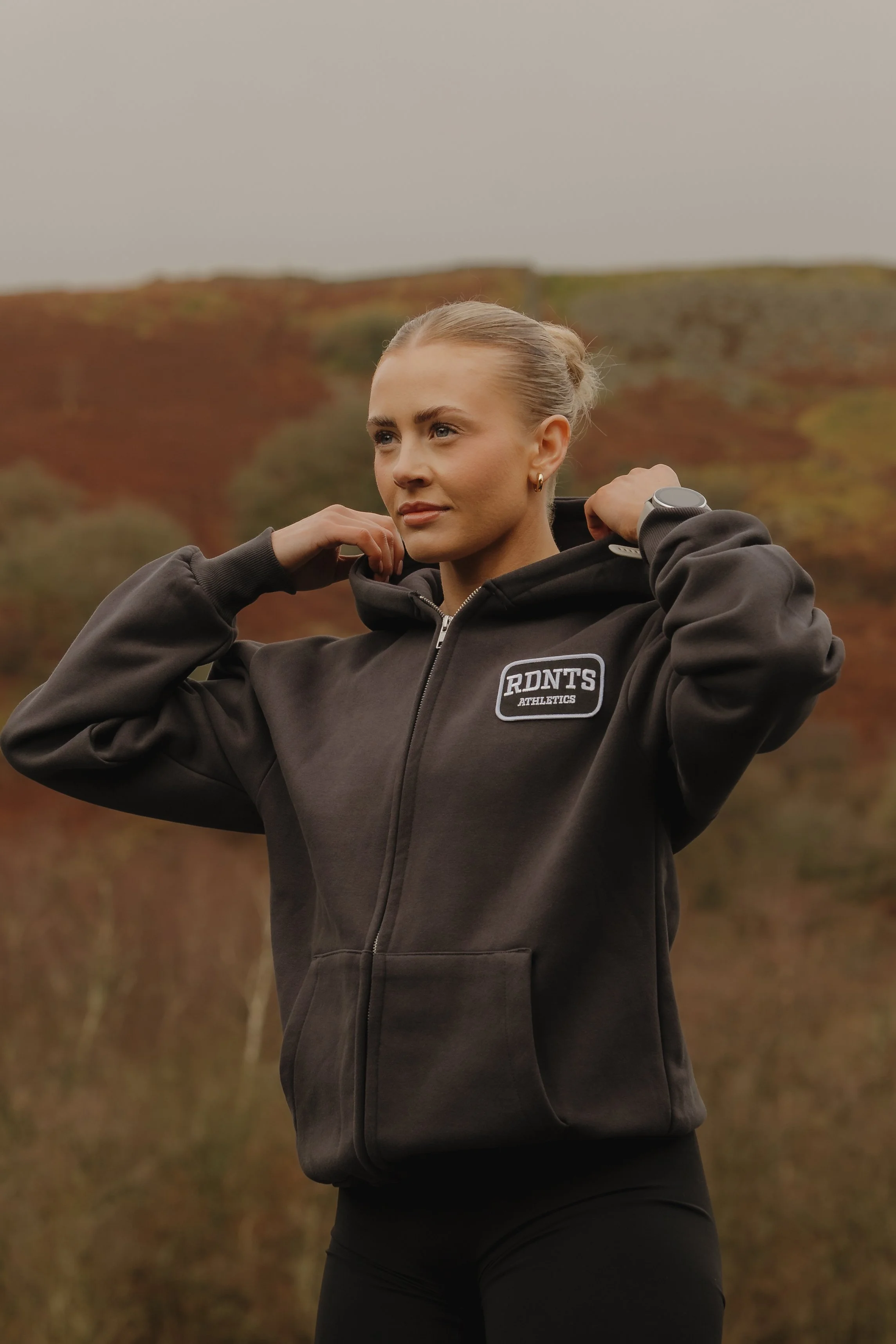 A woman in a dark hoodie with "RDNT'S ATHLETICS" logo, adjusting the hood outdoors against a background of colorful autumn foliage.
