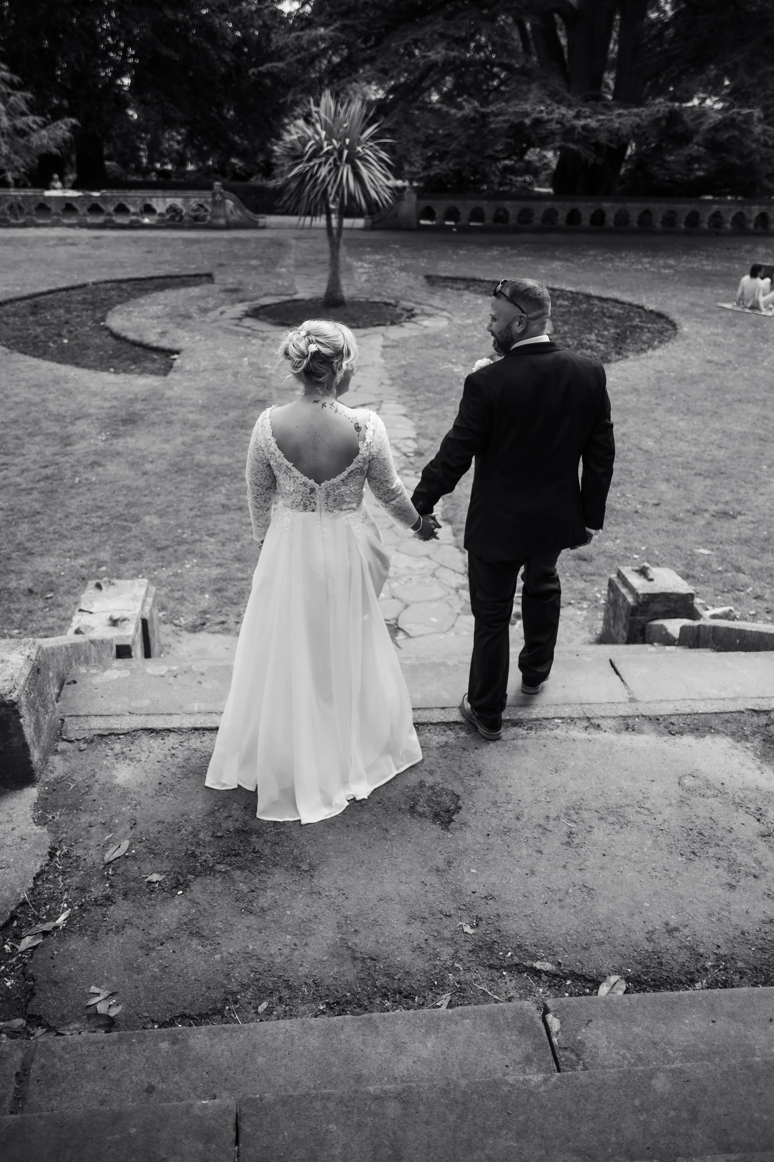 A bride and groom walking hand in hand down steps into a park, viewed from behind. The bride is wearing a long, lace wedding dress with an open back, and the groom is in a dark suit. The park has a stone path and a small palm tree, with trees and a decorative stone wall in the background.