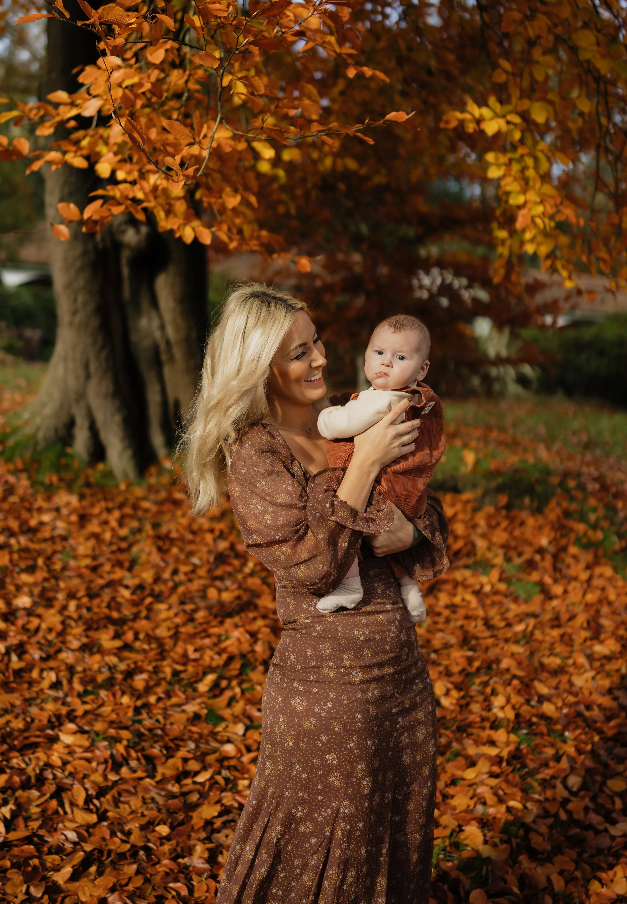 A woman with blonde hair smiling and holding a baby in an autumn park with orange and yellow leaves.