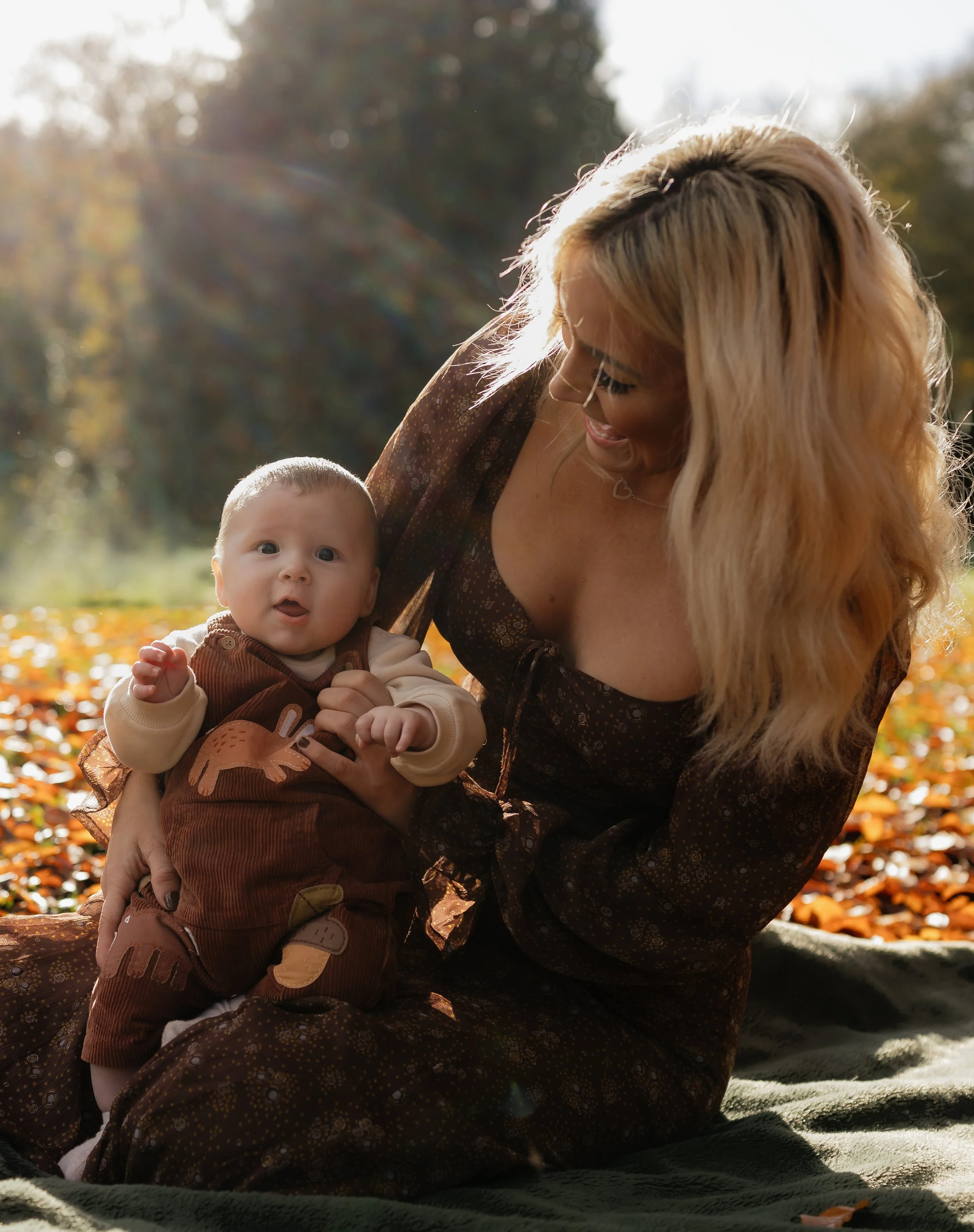 A woman with long blonde hair holding a baby outdoors on autumn foliage, both looking at the camera.