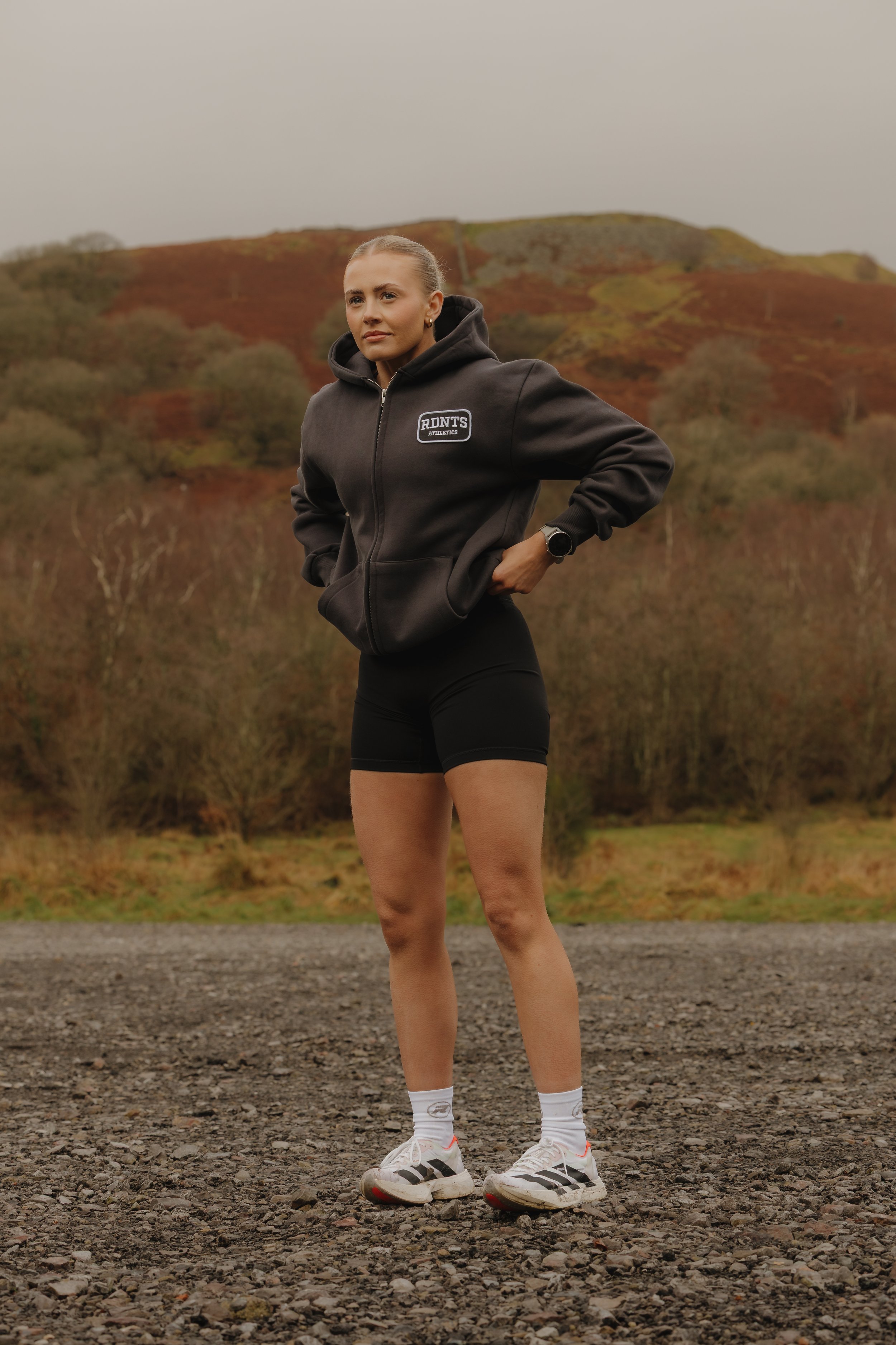 A woman standing outdoors on a gravel surface, wearing a black hoodie, black shorts, white socks, and athletic shoes, with a scenic background of hills and trees.