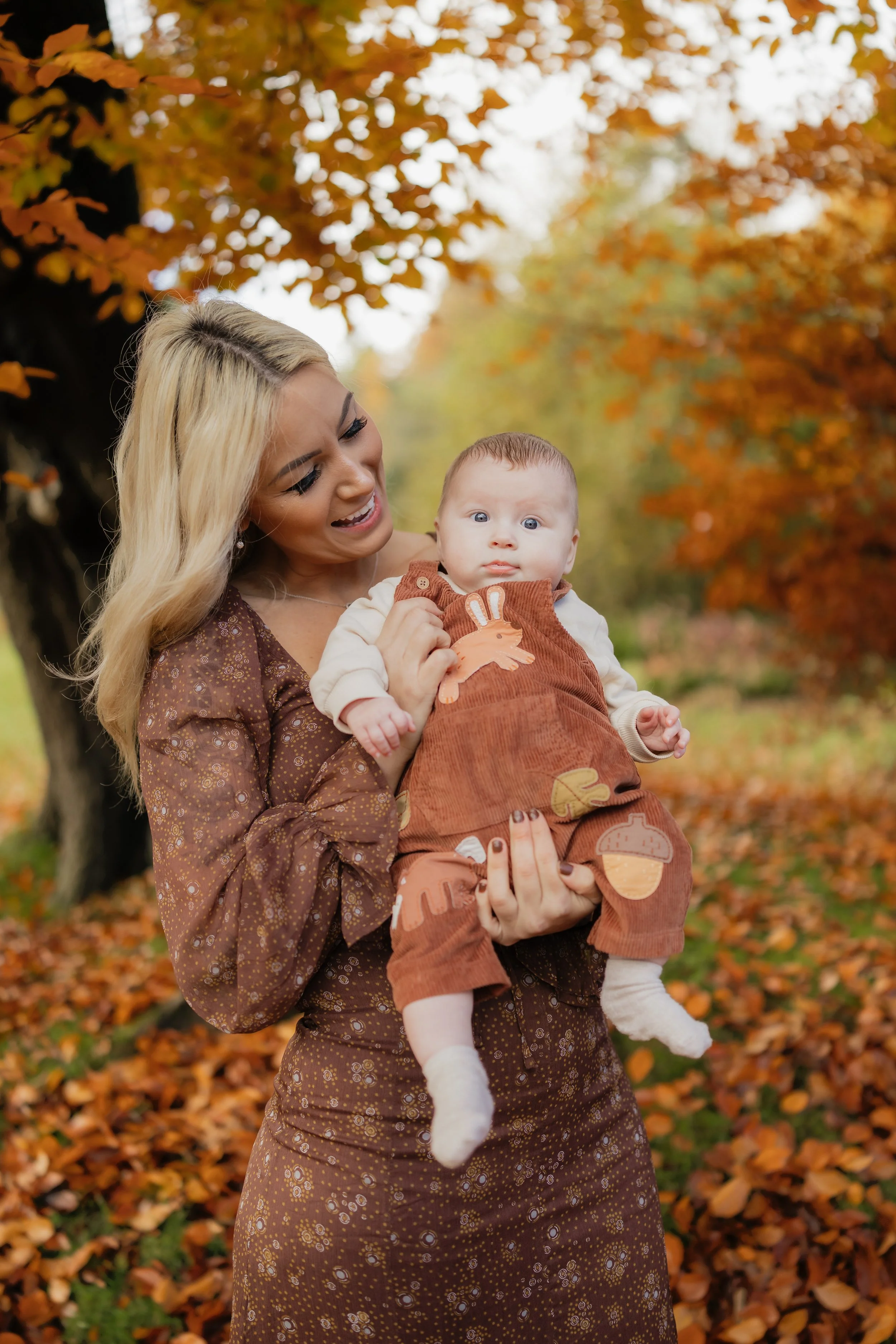 A smiling woman holds a baby in an outdoor park with autumn leaves and trees in the background.