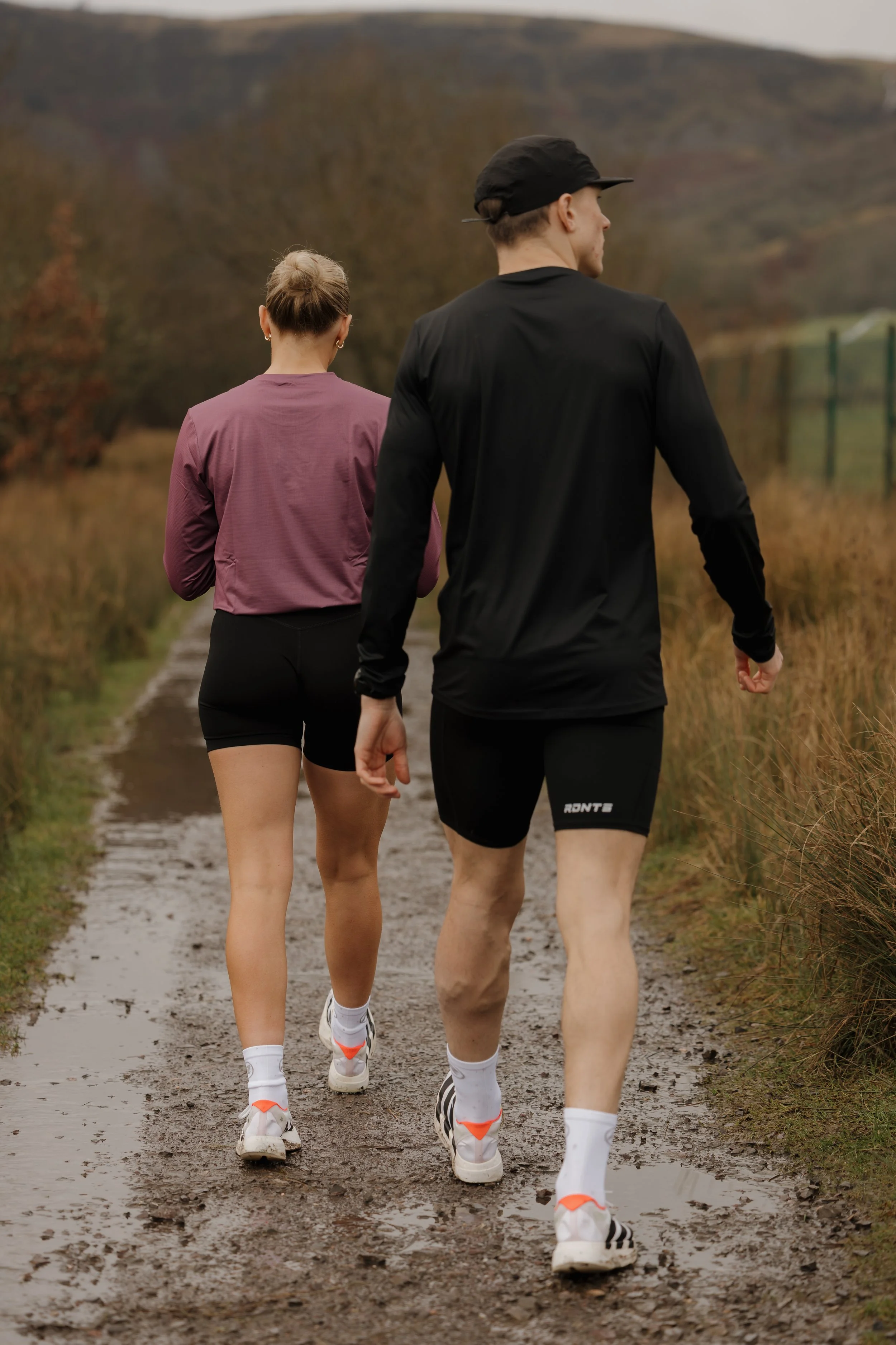 Two people jogging on a muddy trail in a rural area during autumn, with hills in the background and fallen leaves.