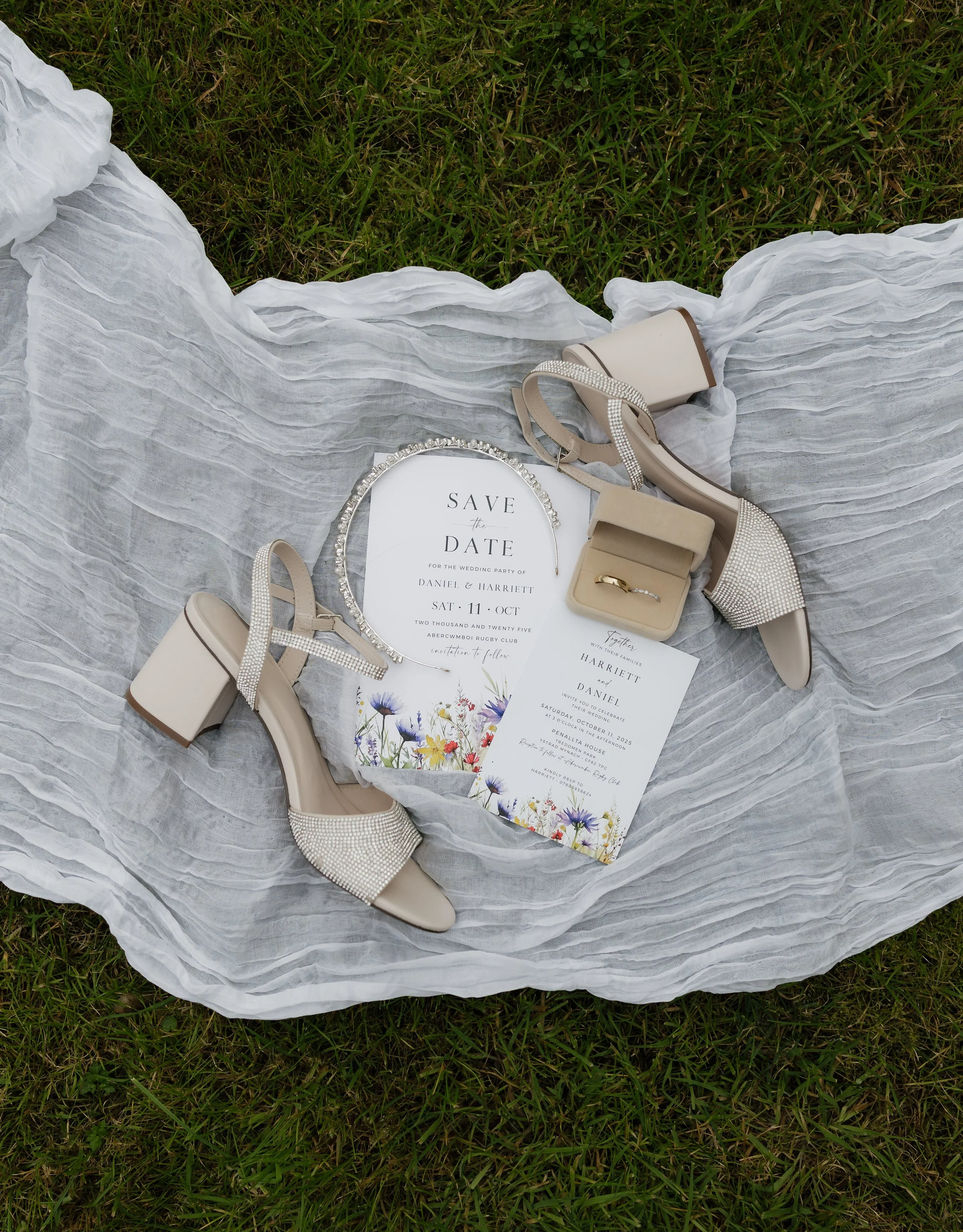 Wedding accessories including white high-heeled shoes with rhinestone details, invitation cards with floral designs, a silver ring, a small beige jewelry box, and a small light-colored purse, all arranged on a white sheer fabric on grass.