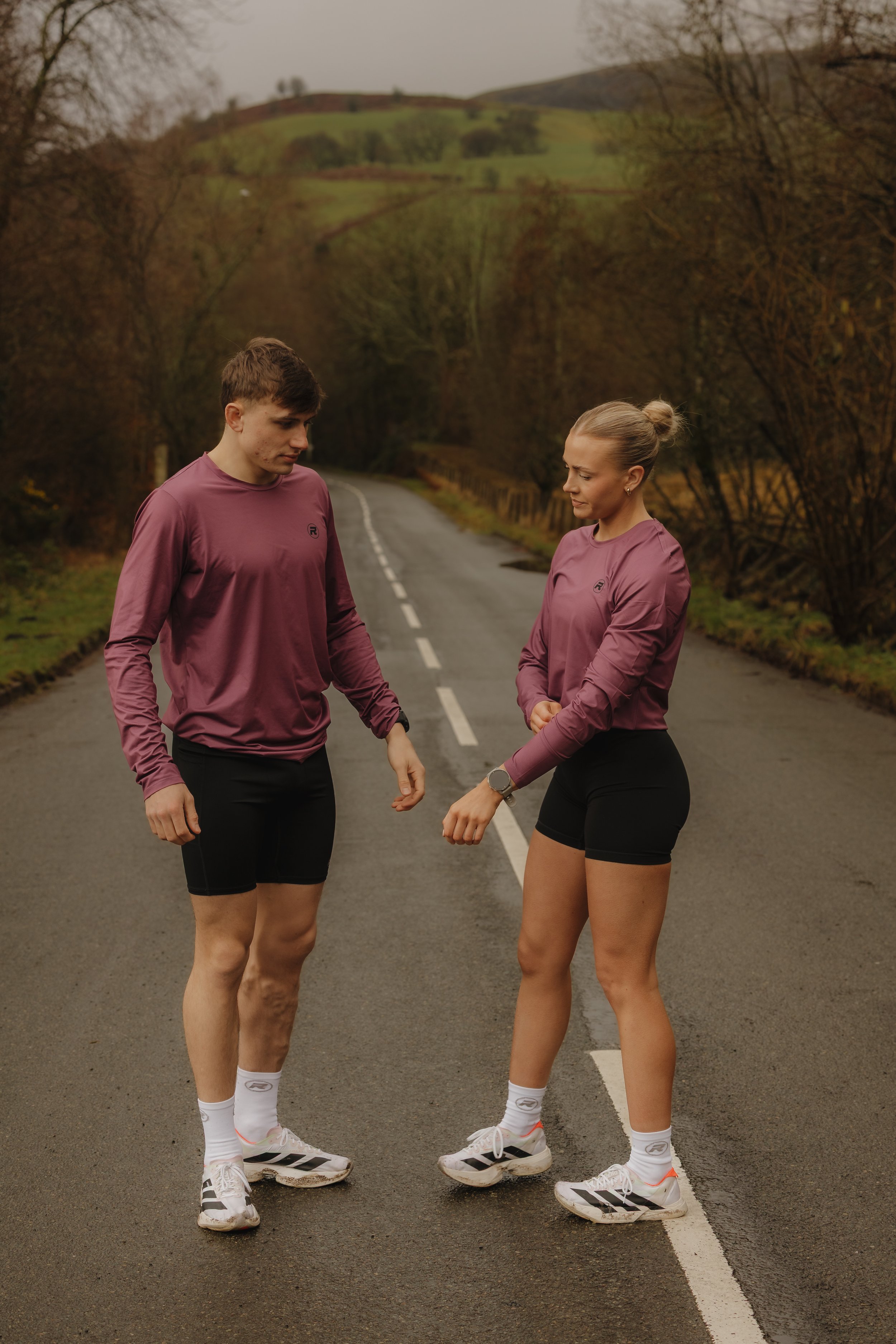 A young man and woman wearing athletic clothing, standing on a deserted rural road, looking at their wrists as if checking a watch, with a scenic background of trees and rolling hills.