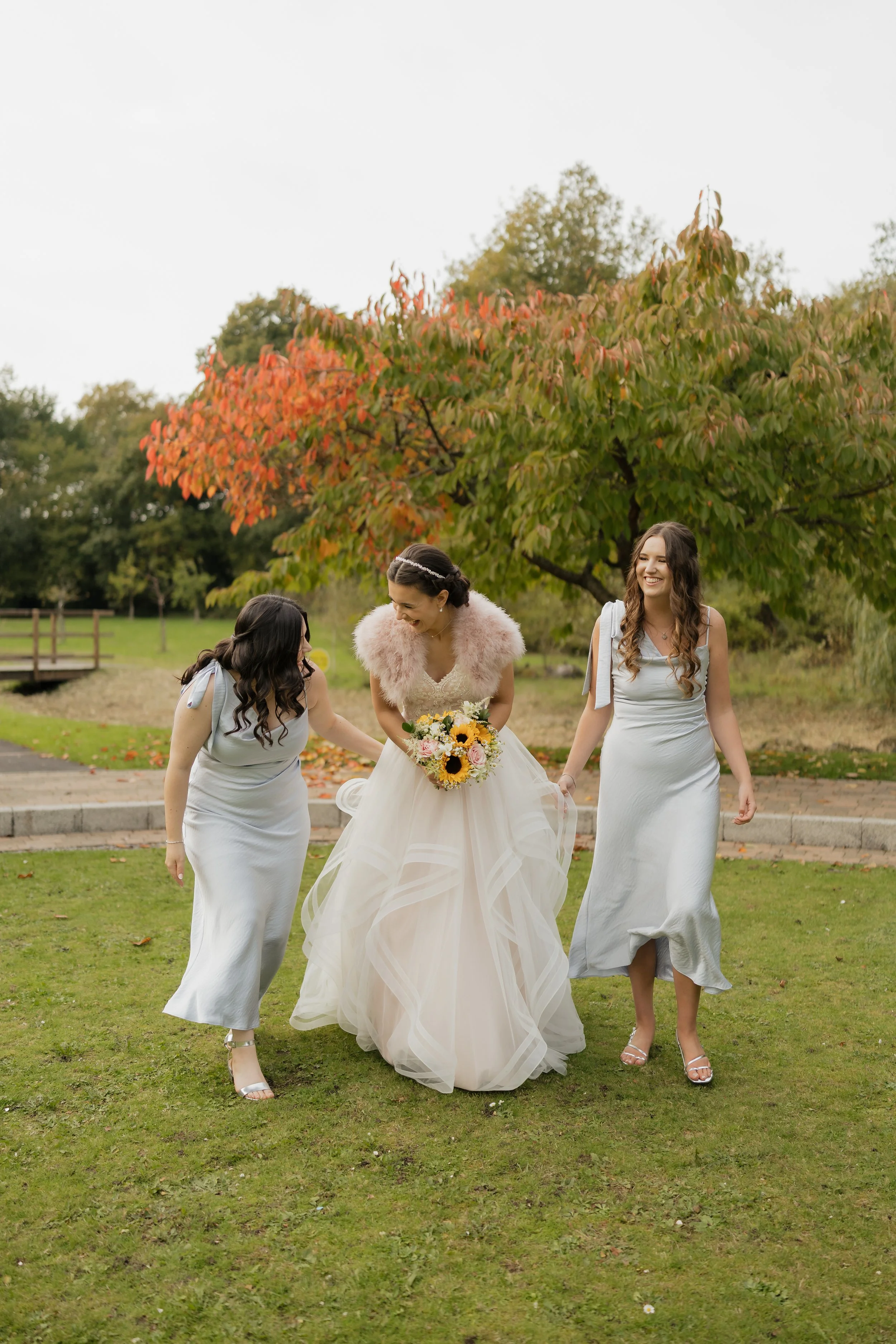 A bride in a gown holding a bouquet of sunflowers and roses is flanked by two bridesmaids in silver dresses as they walk in a park with autumn foliage.