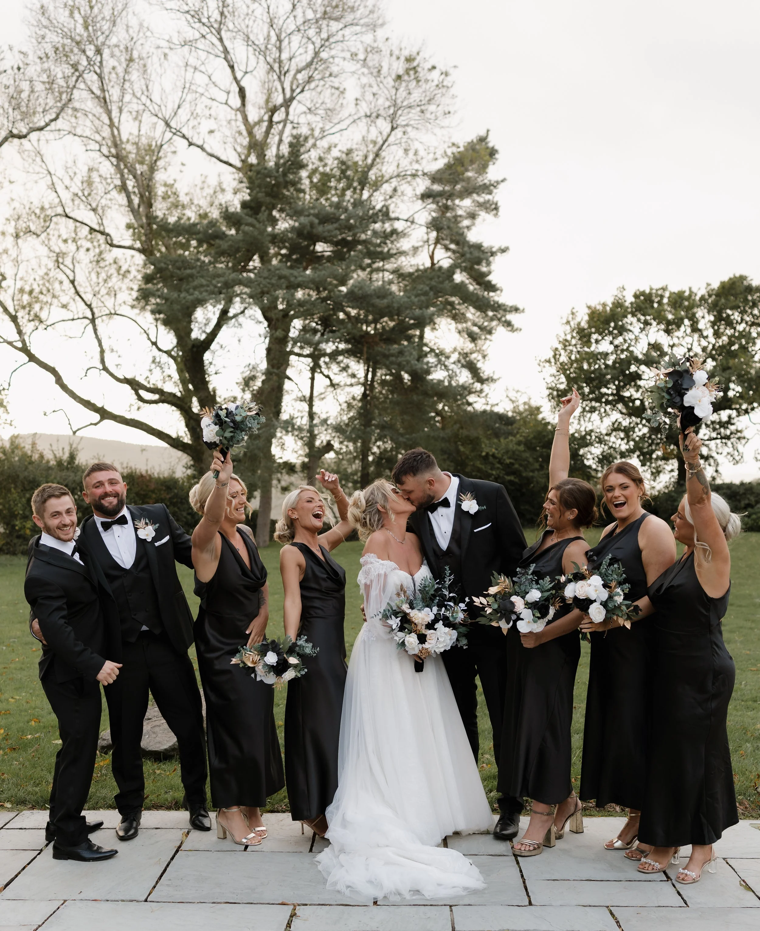 Bride and groom kissing among wedding party outdoors, friends in black dresses and tuxedos celebrating with bouquets.