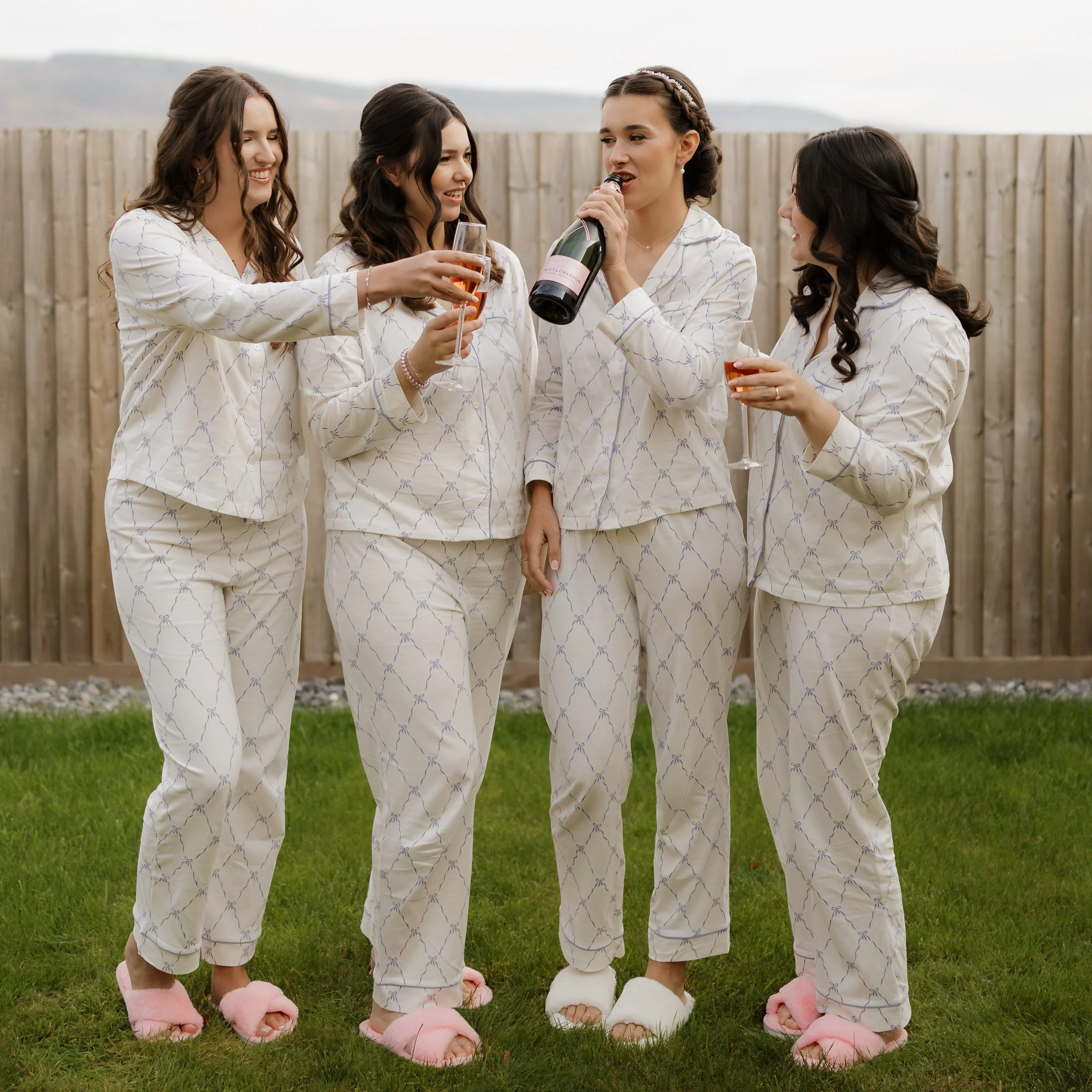 Four women in pajamas and pink slippers celebrating outdoors with champagne, one woman opening a bottle and others holding glasses.