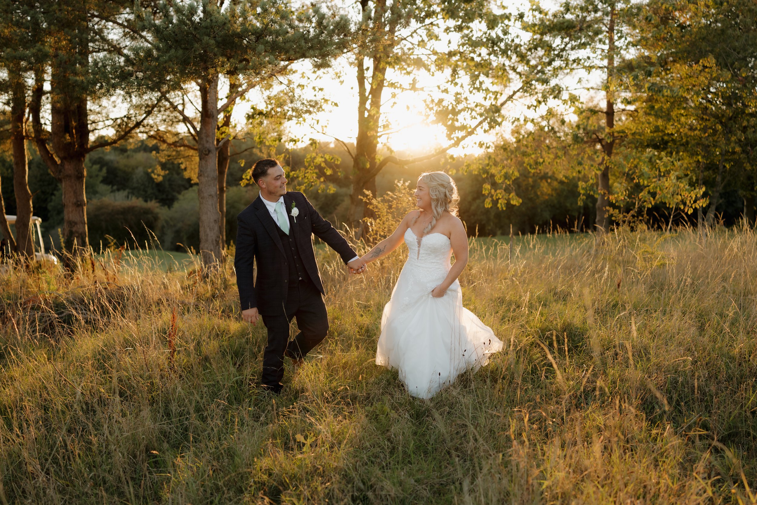A newlywed couple holding hands and smiling at each other in a grassy field during sunset, with trees in the background.