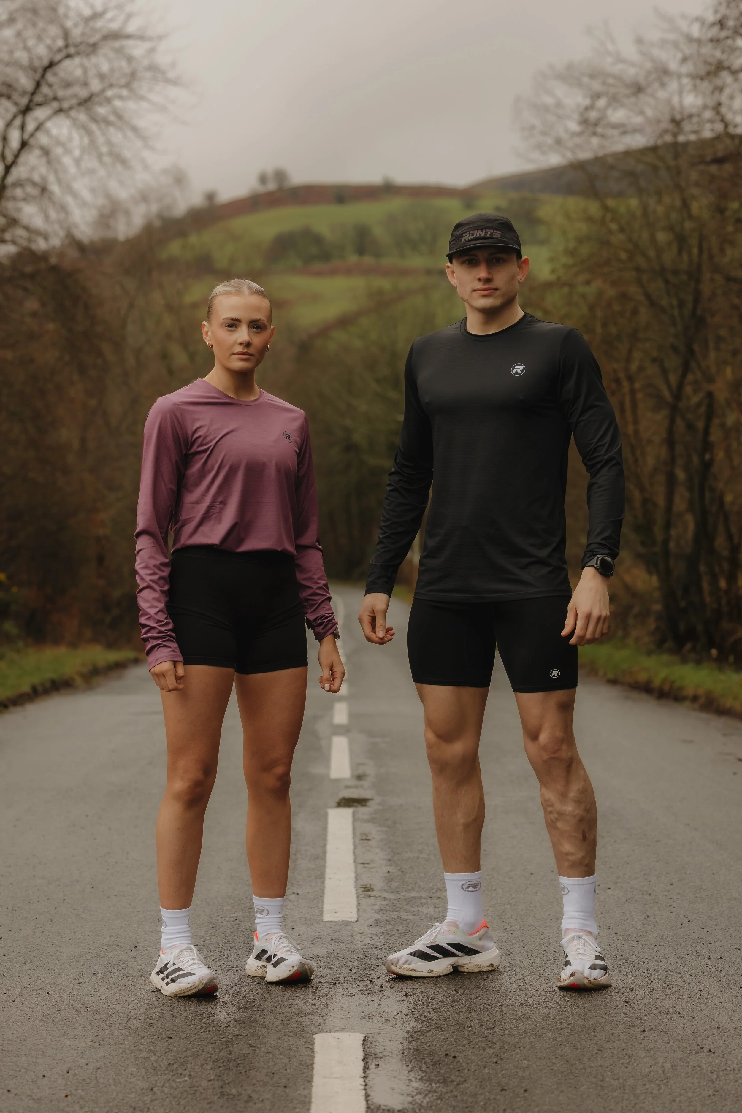 A man and woman standing on a rural road wearing athletic clothing, with autumn trees and hills in the background.