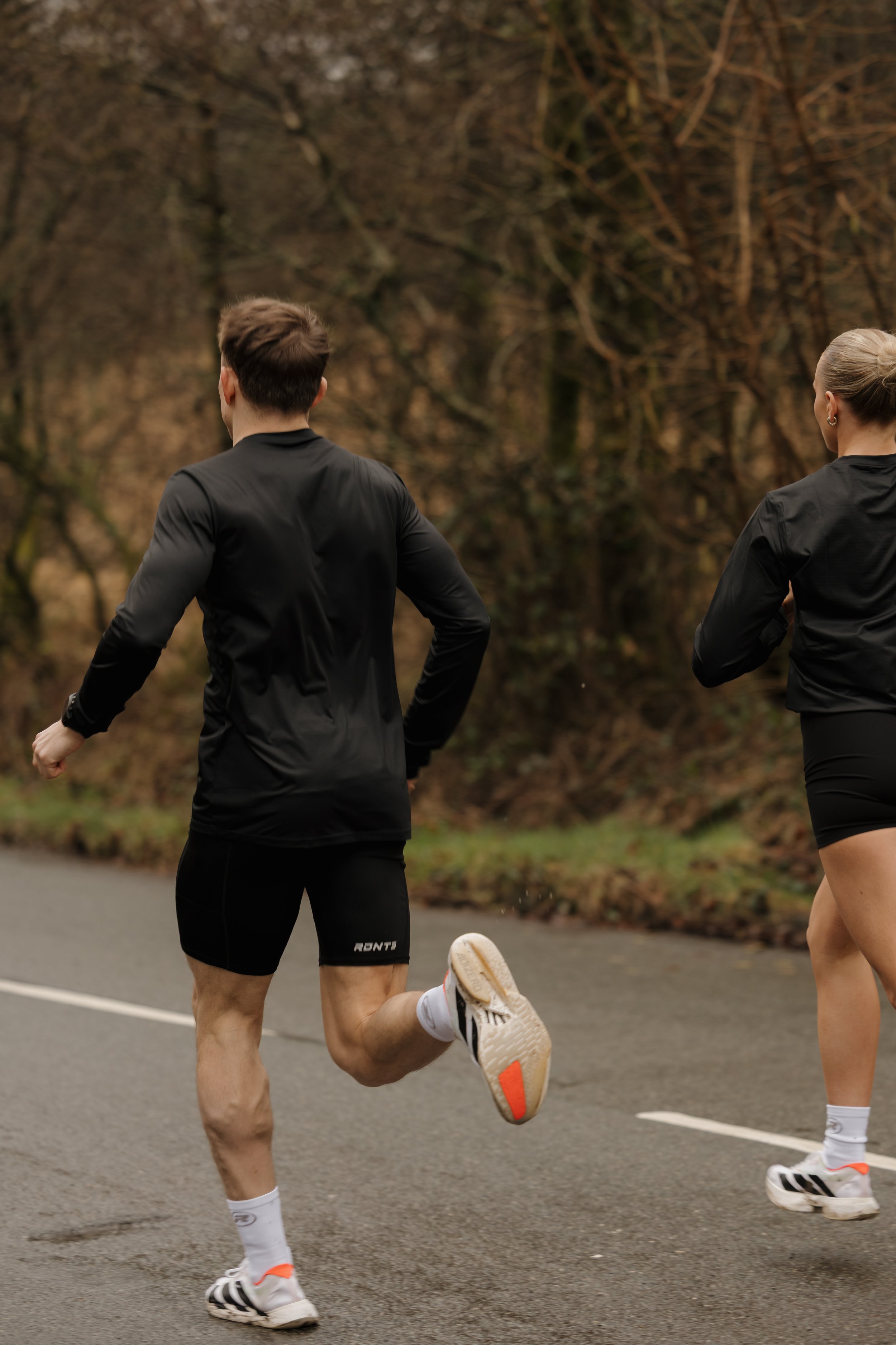 Two people running outdoors on a paved path through a wooded area, dressed in black athletic clothing.