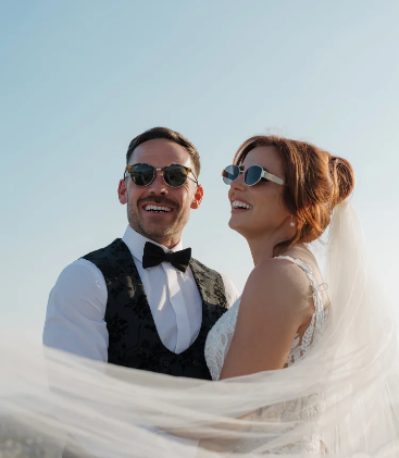 A happy couple at their wedding, the bride and groom are wearing sunglasses and smiling under a flowing veil.