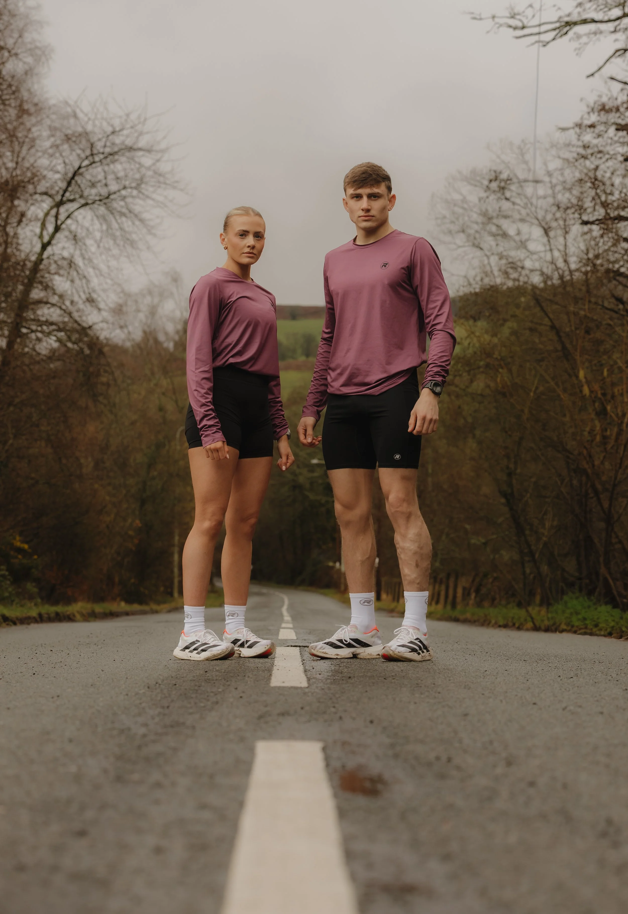 Two young athletes, a woman and a man, standing on a deserted country road, dressed in matching athletic gear, with trees and overcast sky in the background.