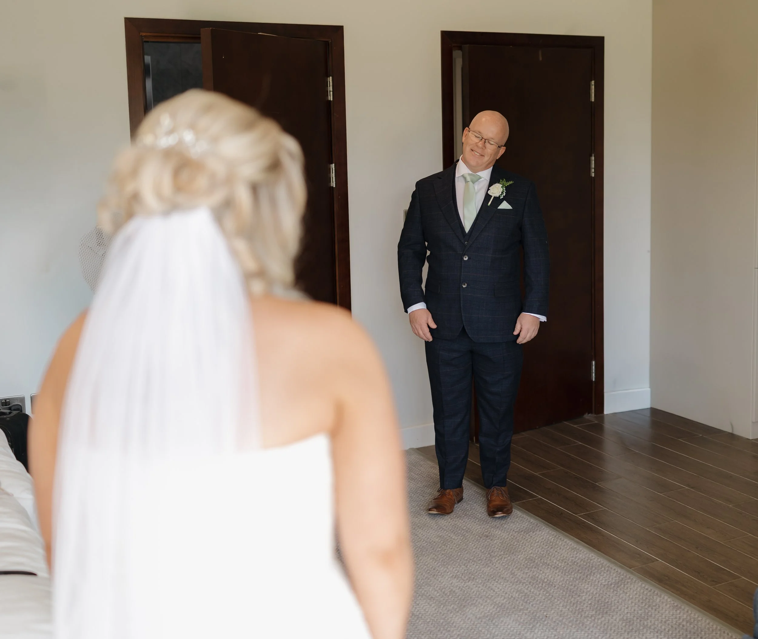 A groom in a dark suit and glasses is smiling as he sees his bride in a white wedding dress with a veil, in a room with wooden floors and white walls.