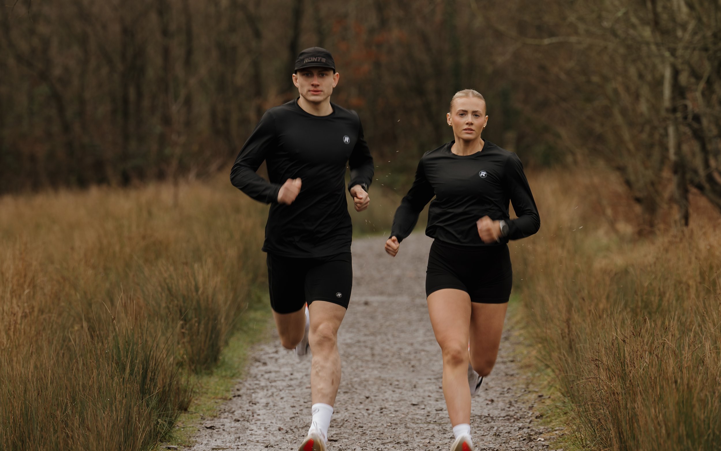 A man and woman jogging on a dirt trail through a natural landscape with trees and grass during autumn.