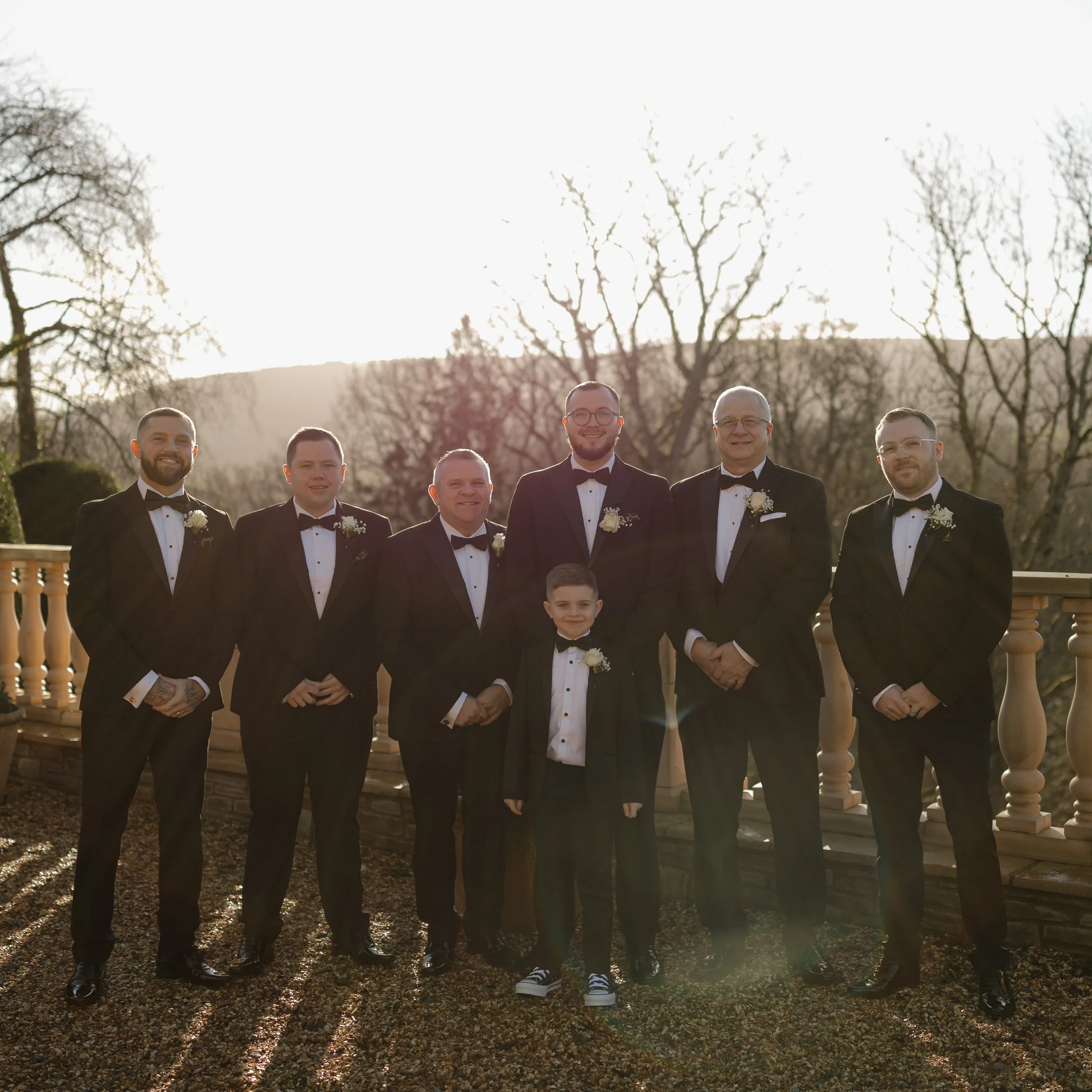 A group of seven men, including a young boy, dressed in tuxedos standing outdoors with trees and a stone railing in the background during sunset.
