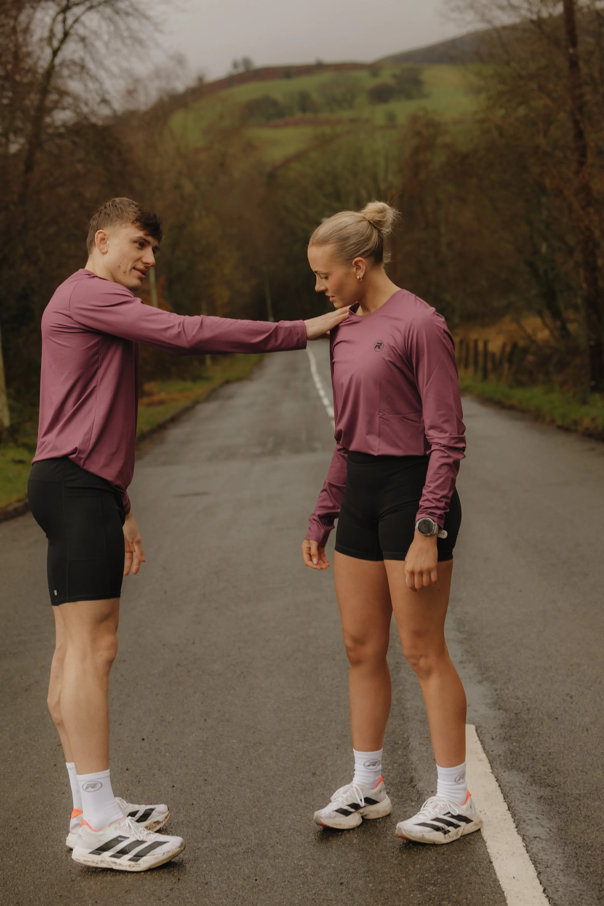 A man with a lean build and a woman with an athletic build dressed in running gear standing on a road in a rural area with trees and rolling hills in the background. The man is touching the woman's shoulder and they appear to be preparing for a run.