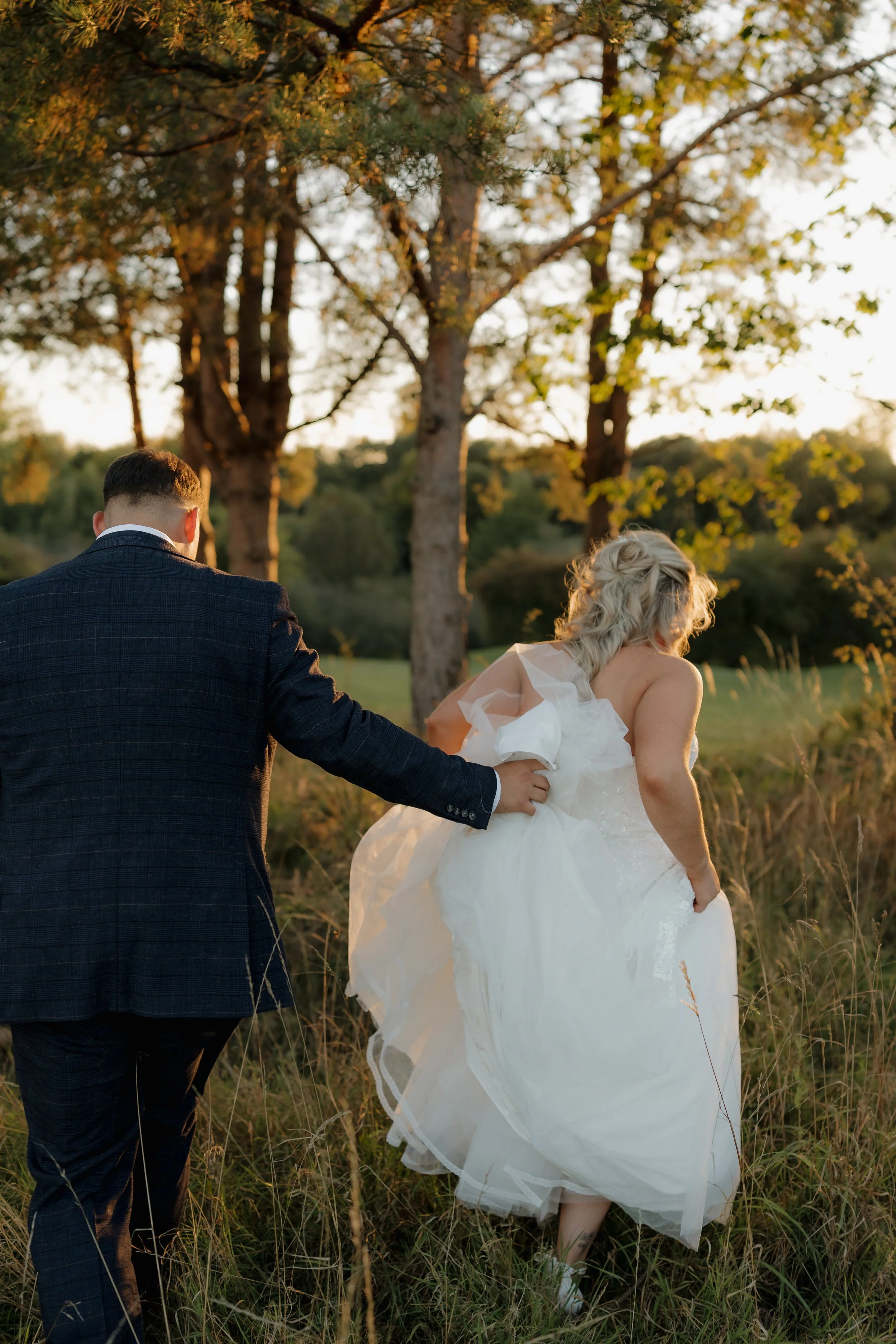 A bride in a white lace wedding dress stands on a grassy field, with a groom in a blue suit walking toward her in the background, on a sunny day with trees in the distance.