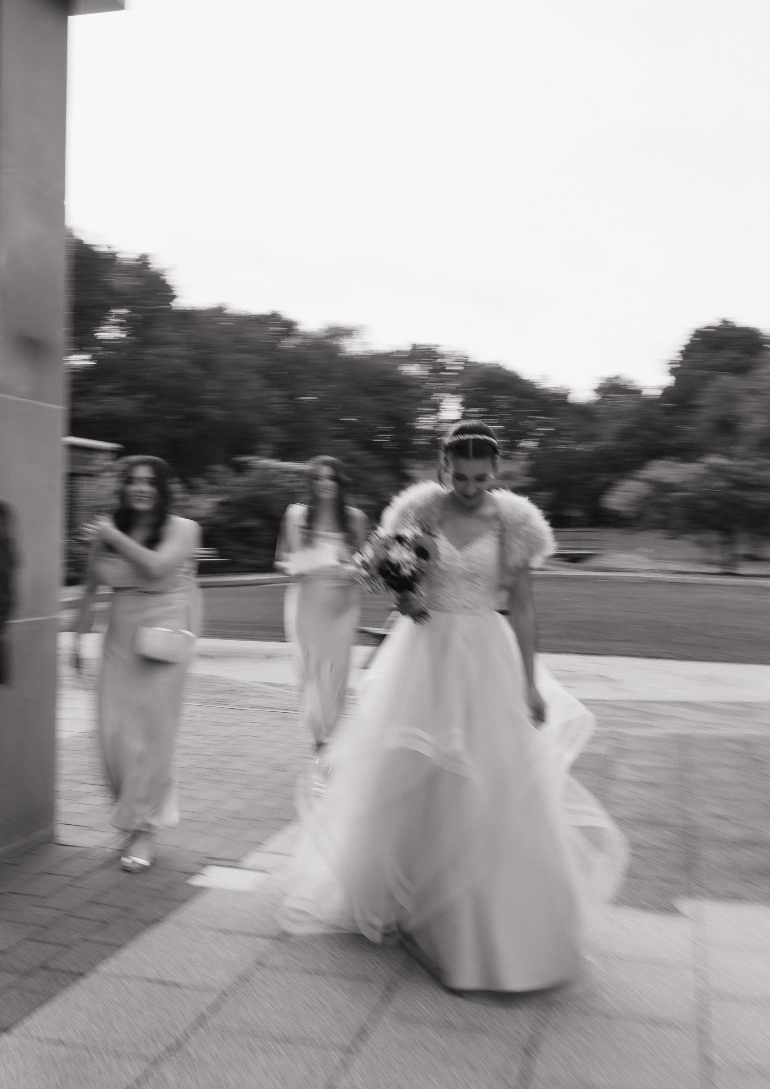 A bride in a wedding dress and fur stole walking outside with bridesmaids in long dresses amid trees.