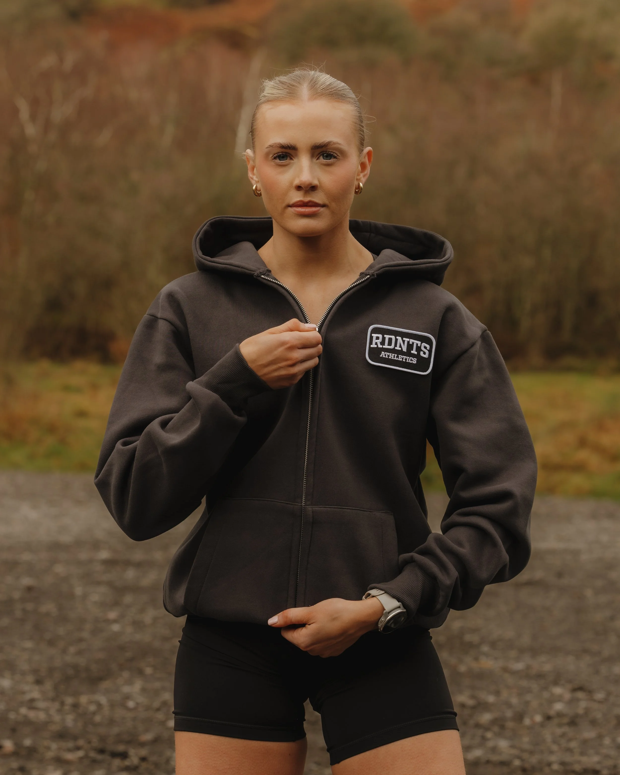 A young woman in athletic clothing standing outdoors on a gravel path with trees in the background. She is wearing a black hoodie with a patch that reads 'RDNT'S Athletics' and black shorts.
