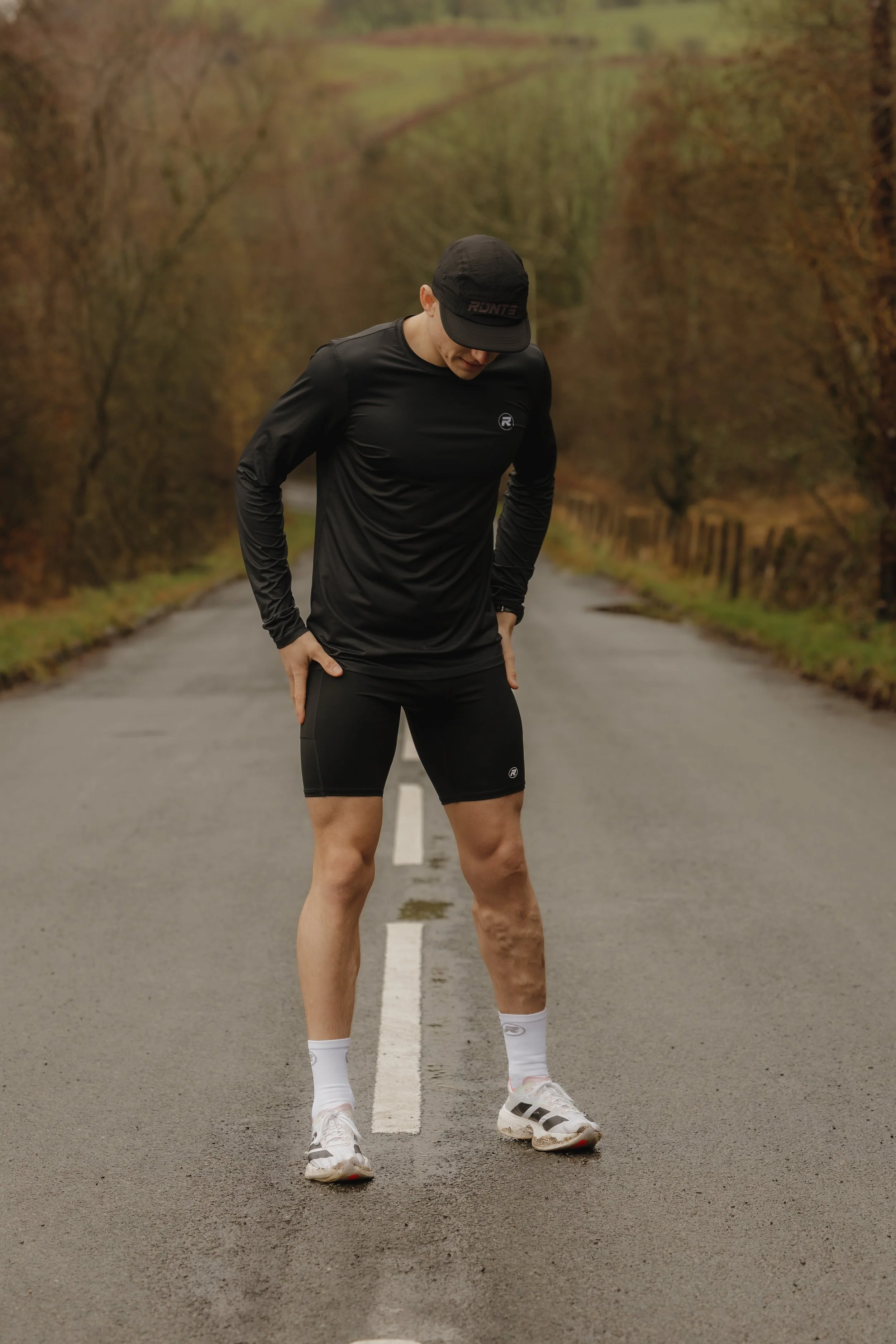 A man dressed in black athletic clothing standing on a wet road, looking down at his legs with a concerned or frustrated expression.