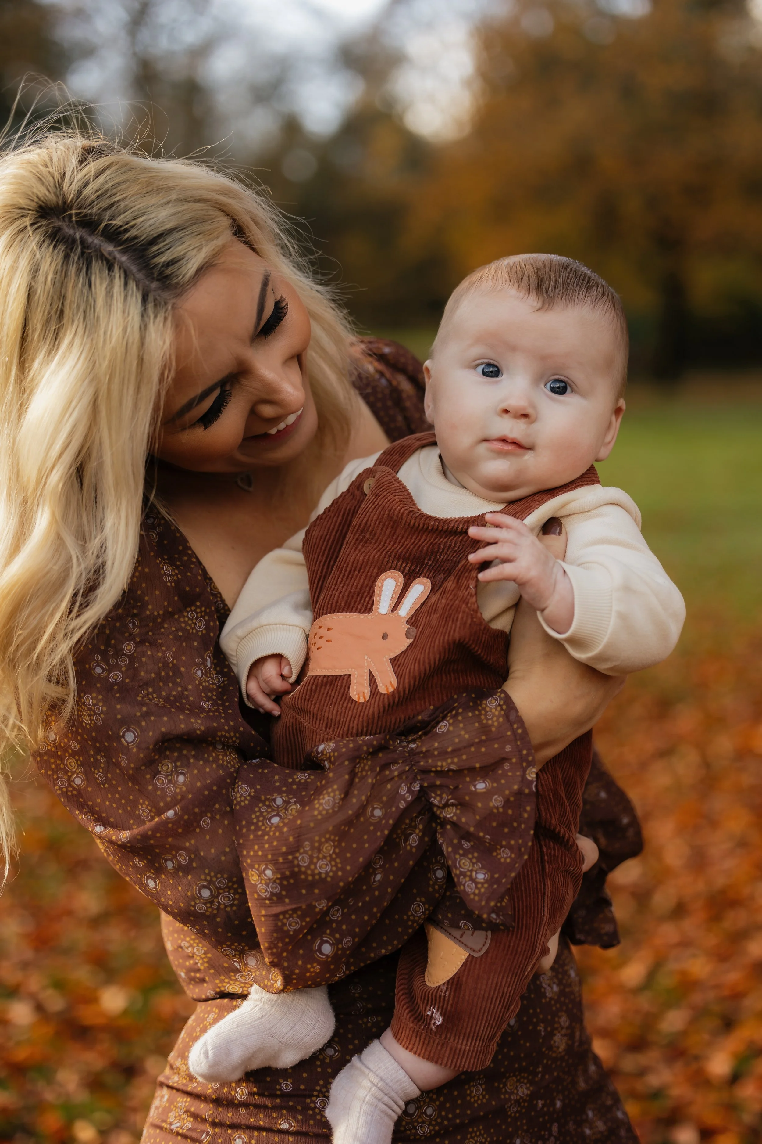 A woman holding a baby outdoors in an autumn setting.