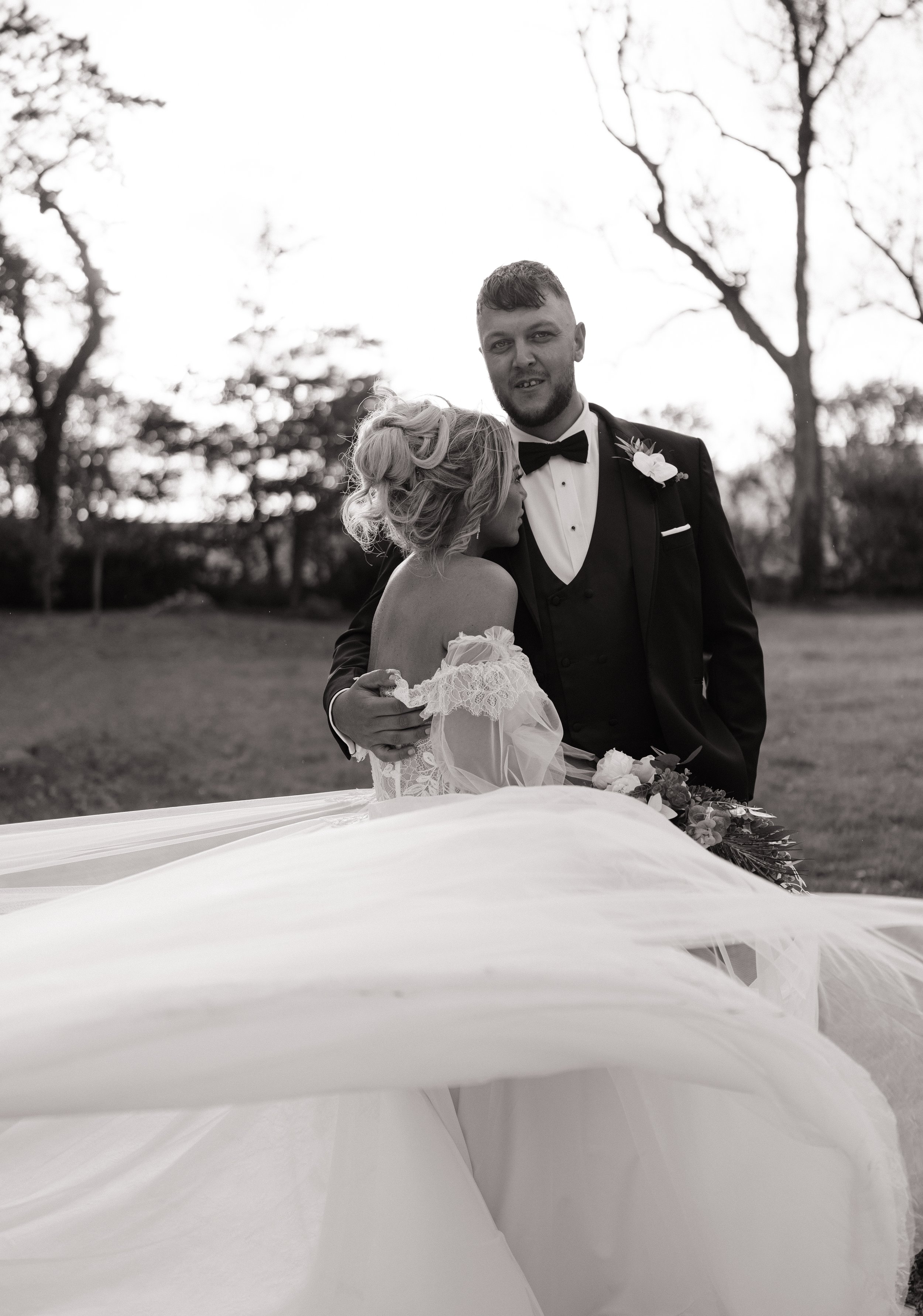 Black and white photo of a bride and groom outdoors, with trees in the background. The bride has blonde hair styled in loose curls, and the groom has short hair and a beard. The groom is wearing a tuxedo with a bow tie and boutonniere, and the bride 
