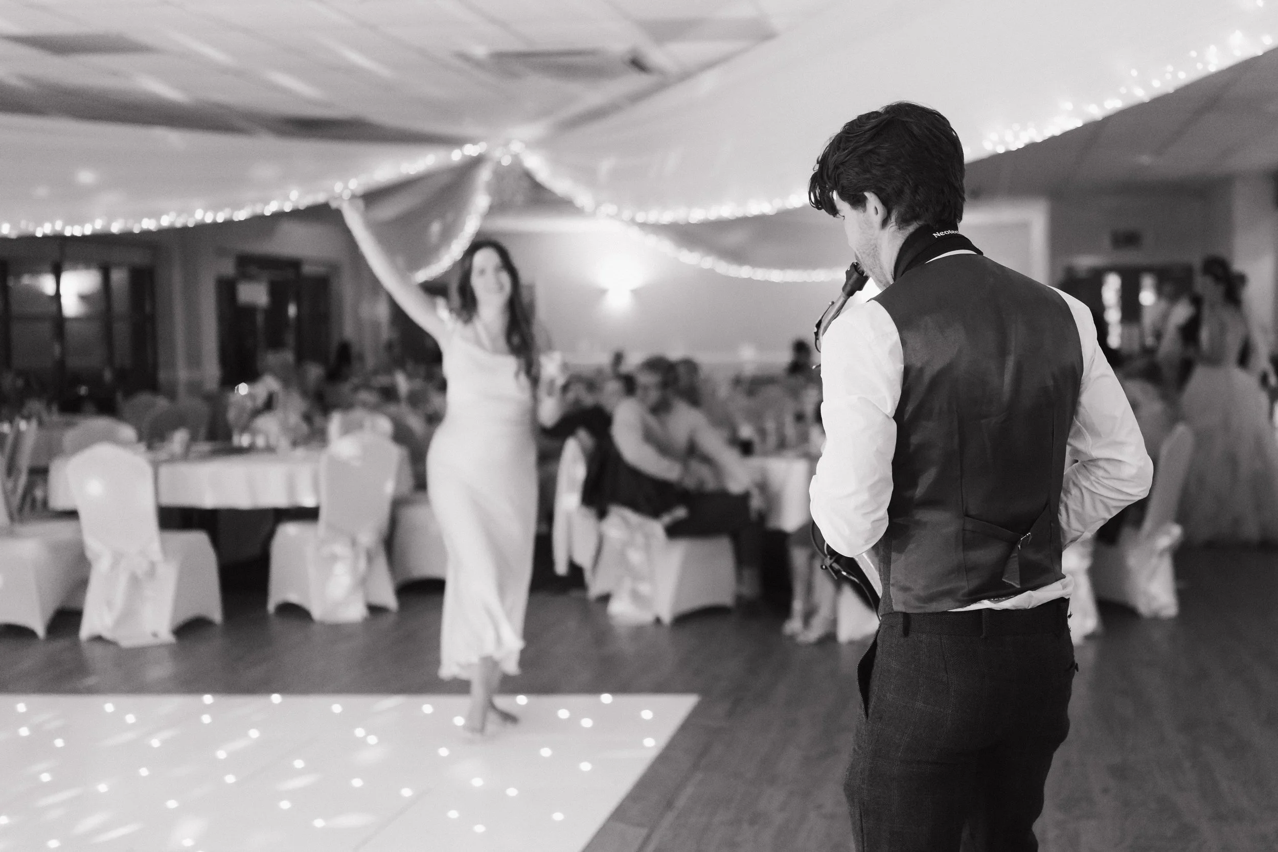 A man in formal attire holding a clarinet while watching a woman in a white dress dancing at a wedding reception with guests sitting at decorated tables in the background.