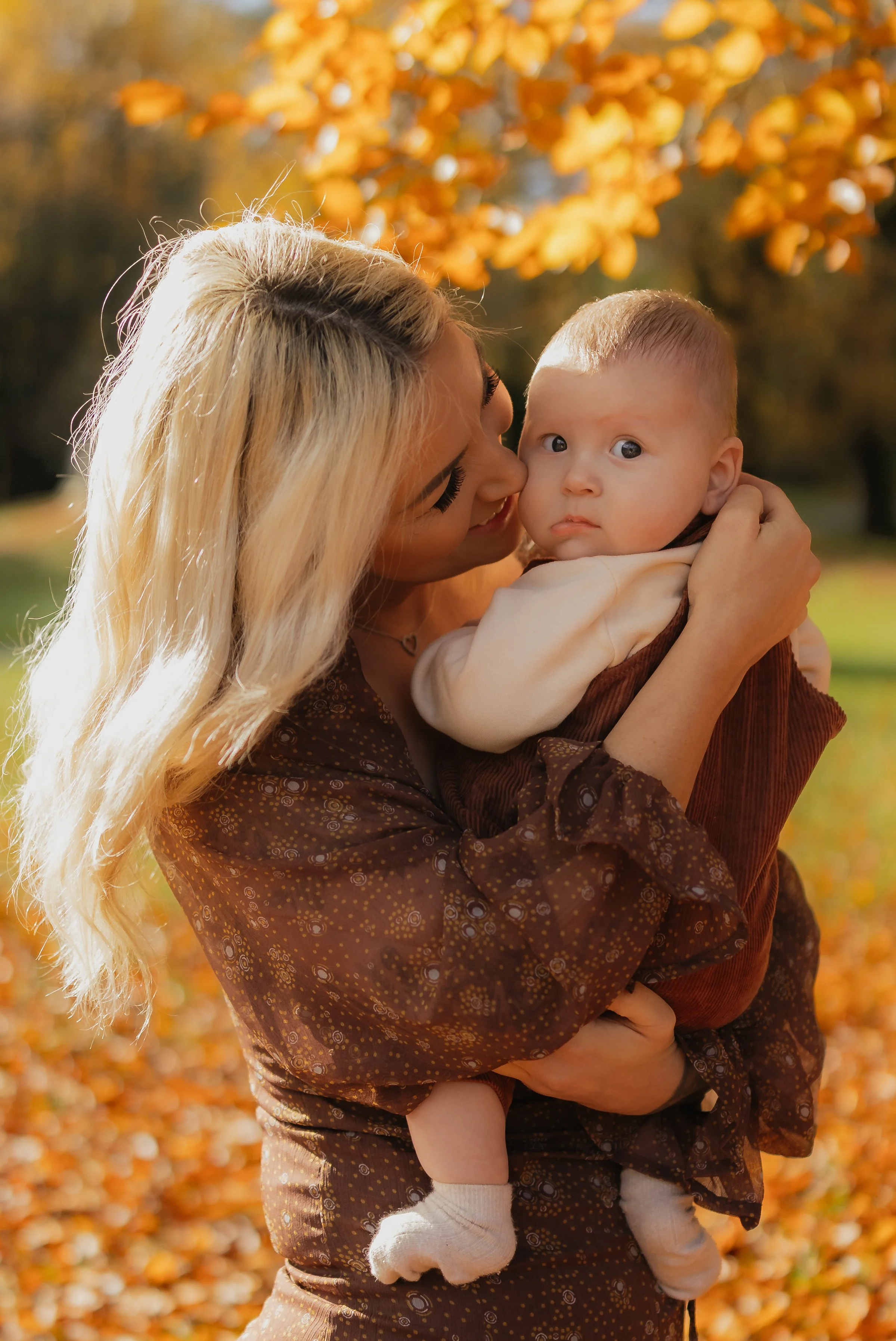 A woman with long blonde hair holding a baby outdoors in fall, with orange leaves in the background.