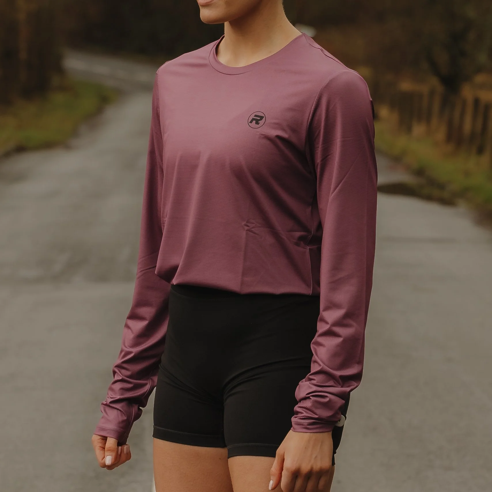 A woman wearing a maroon long-sleeve athletic shirt and black shorts standing outdoors on a paved path surrounded by trees.