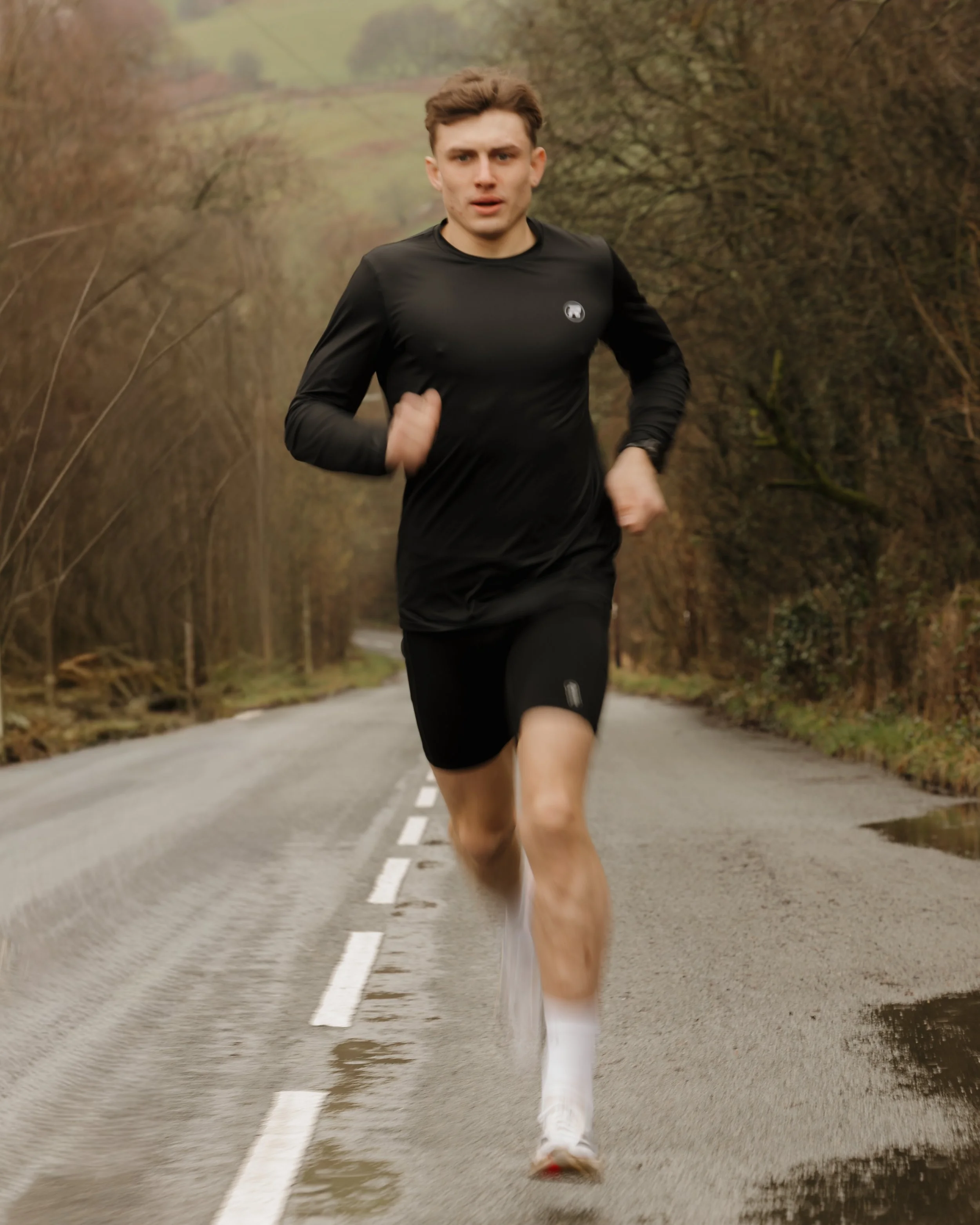 A young man running on a rural road through a wooded area, wearing black athletic clothing and white running shoes.