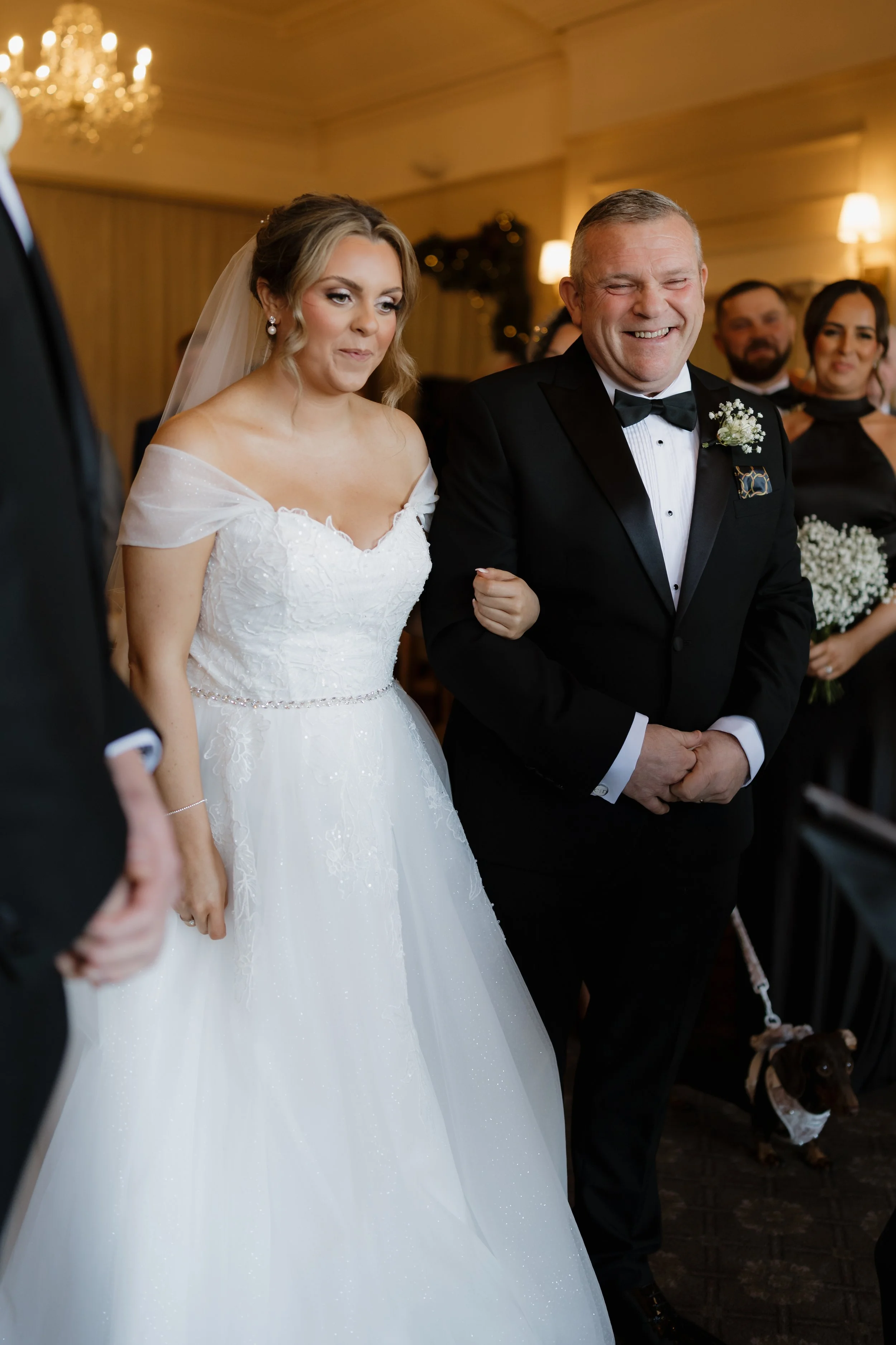 A bride in a white wedding gown with lace details stands next to a smiling man in a tuxedo at a wedding ceremony. In the background, wedding guests are visible, and a small dog on a leash is at the groom's feet.