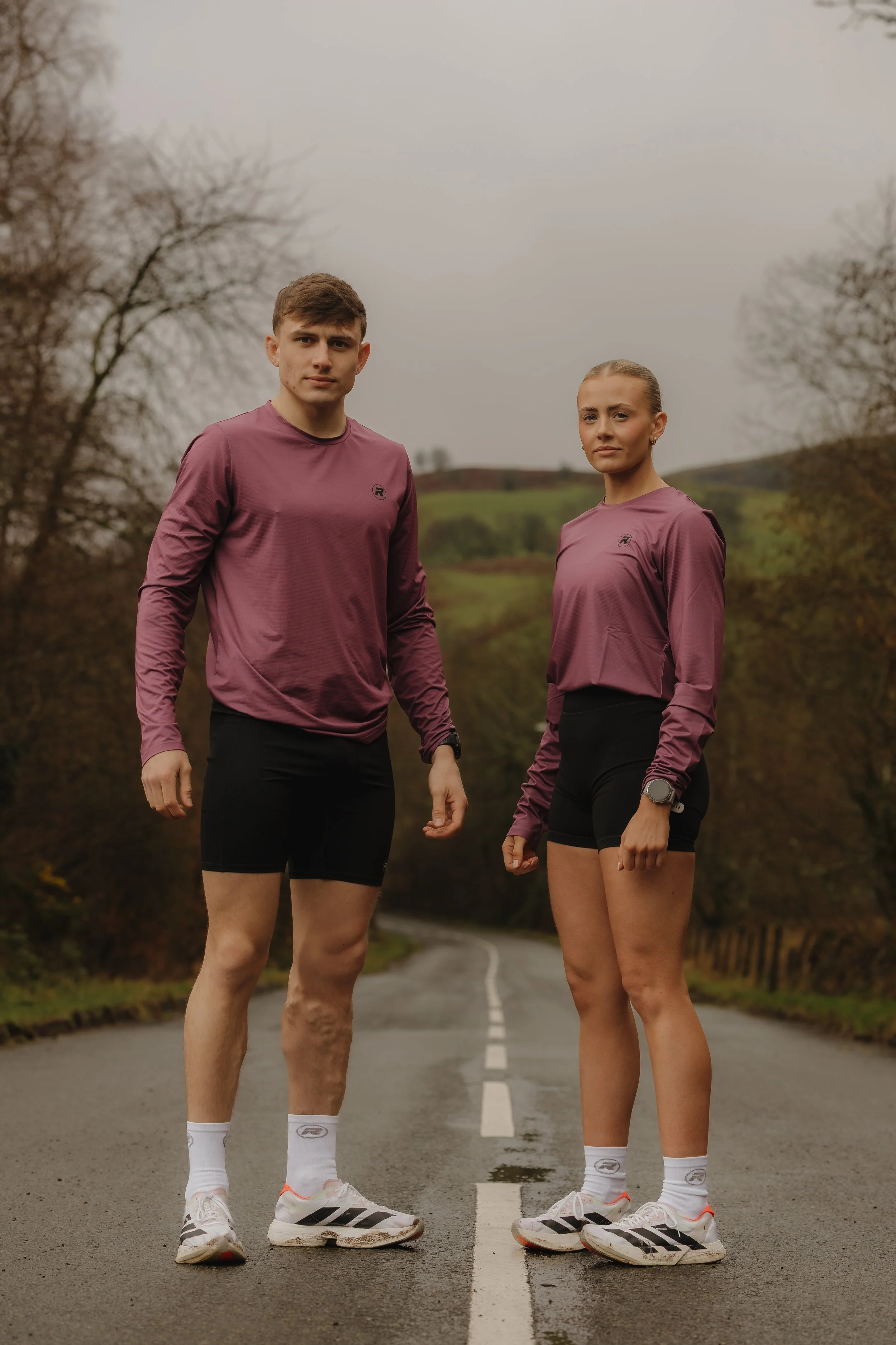 Two young athletes, a man and a woman, standing on a rural road, dressed in matching athletic wear, with an overcast sky and bare trees in the background.