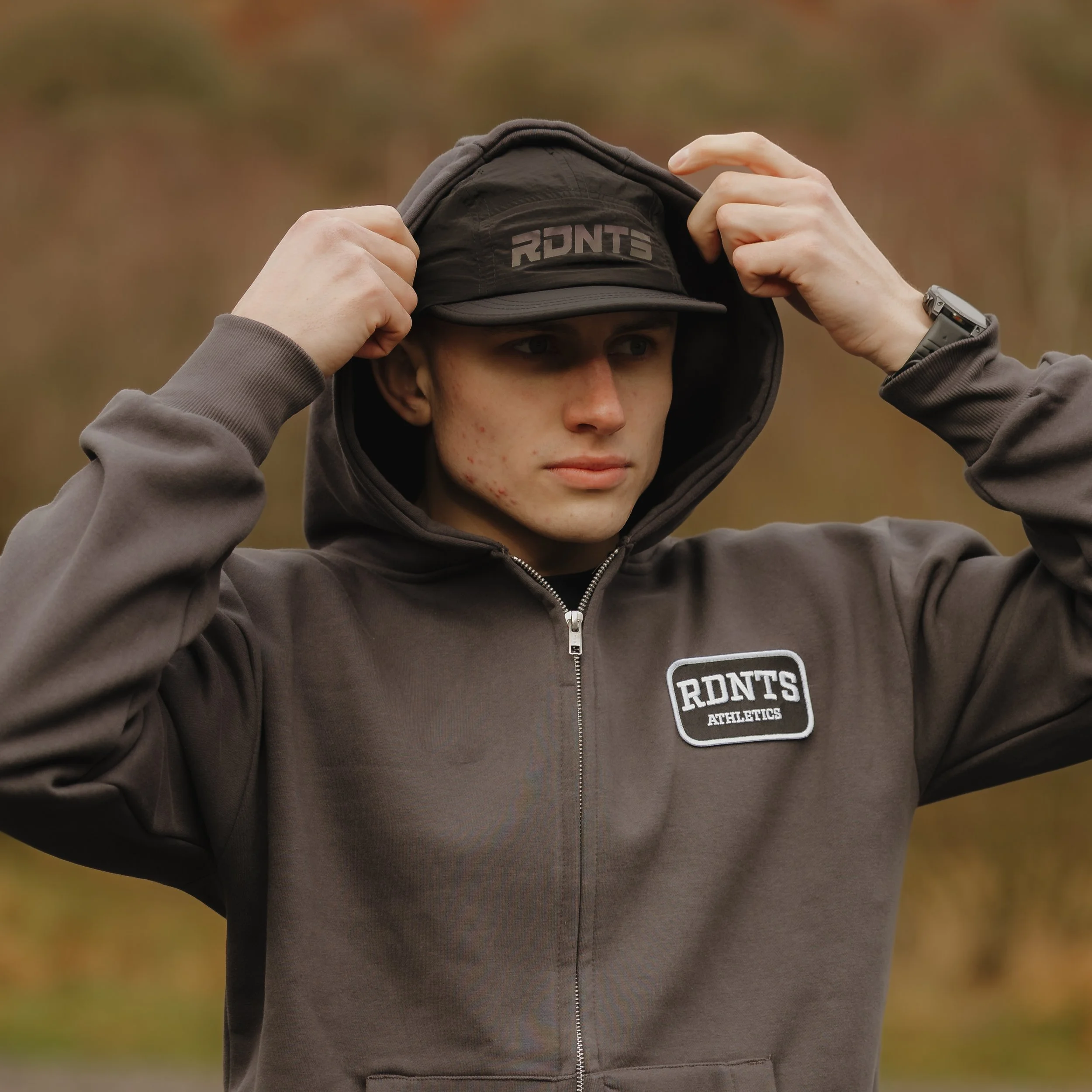 A young man adjusts his hood outdoors, wearing a gray zip-up jacket with a patch that reads 'RDNT'S ATHLETICS' and a black cap with the same text. He has a serious expression, and the background is blurred with autumn colors.