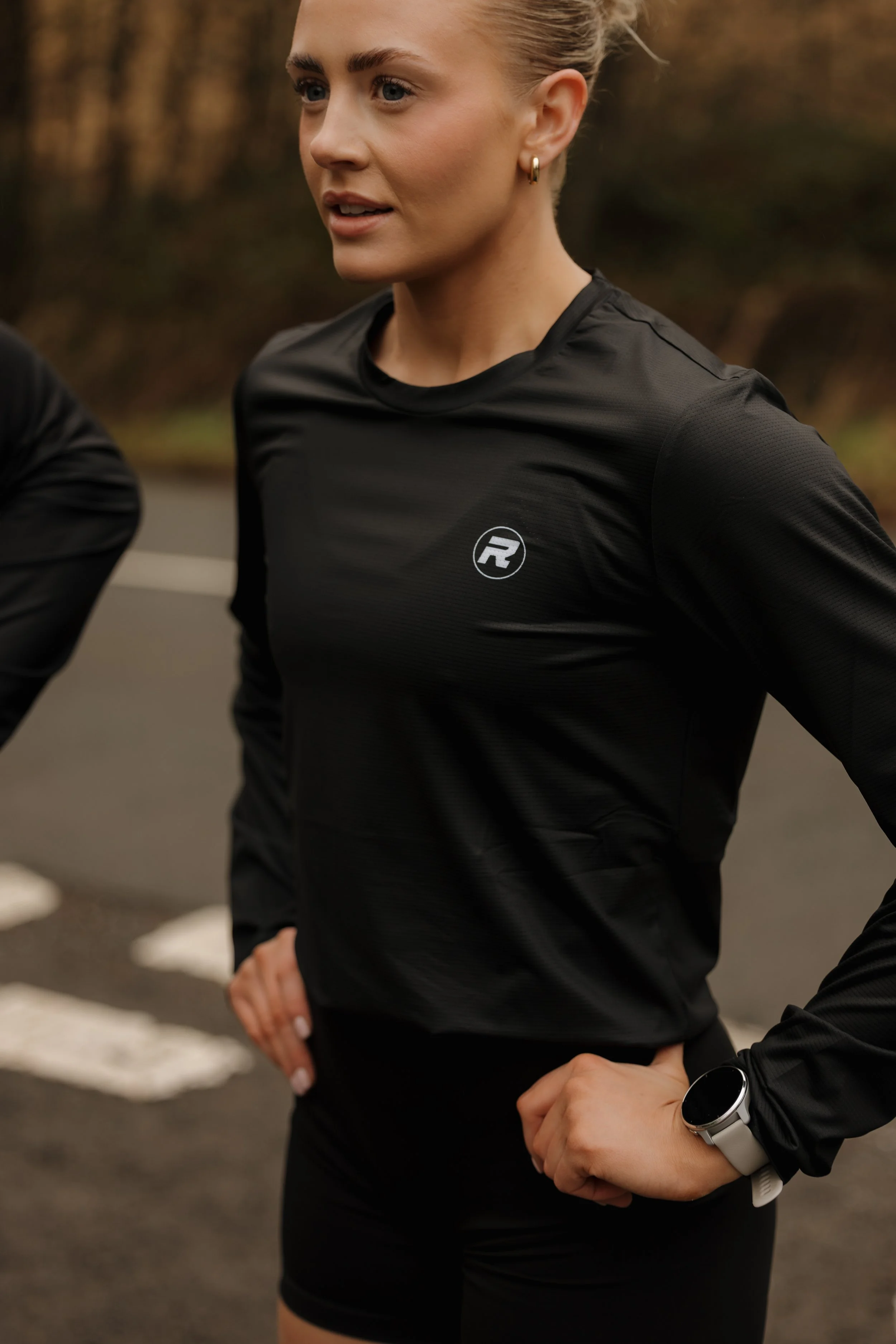A woman with blonde hair in a ponytail wearing a black athletic long-sleeve shirt with a logo, black shorts, and a smartwatch on her left wrist, standing outdoors with her hands on her hips.