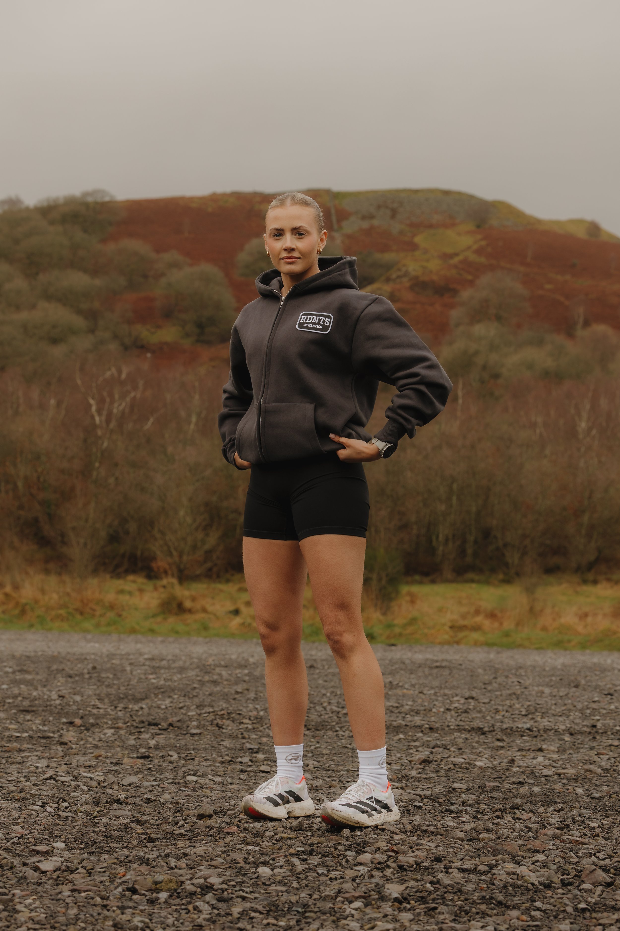 A woman standing outdoors on a gravel field with hills and trees in the background, wearing athletic clothing including a black hoodie, black shorts, and running shoes.