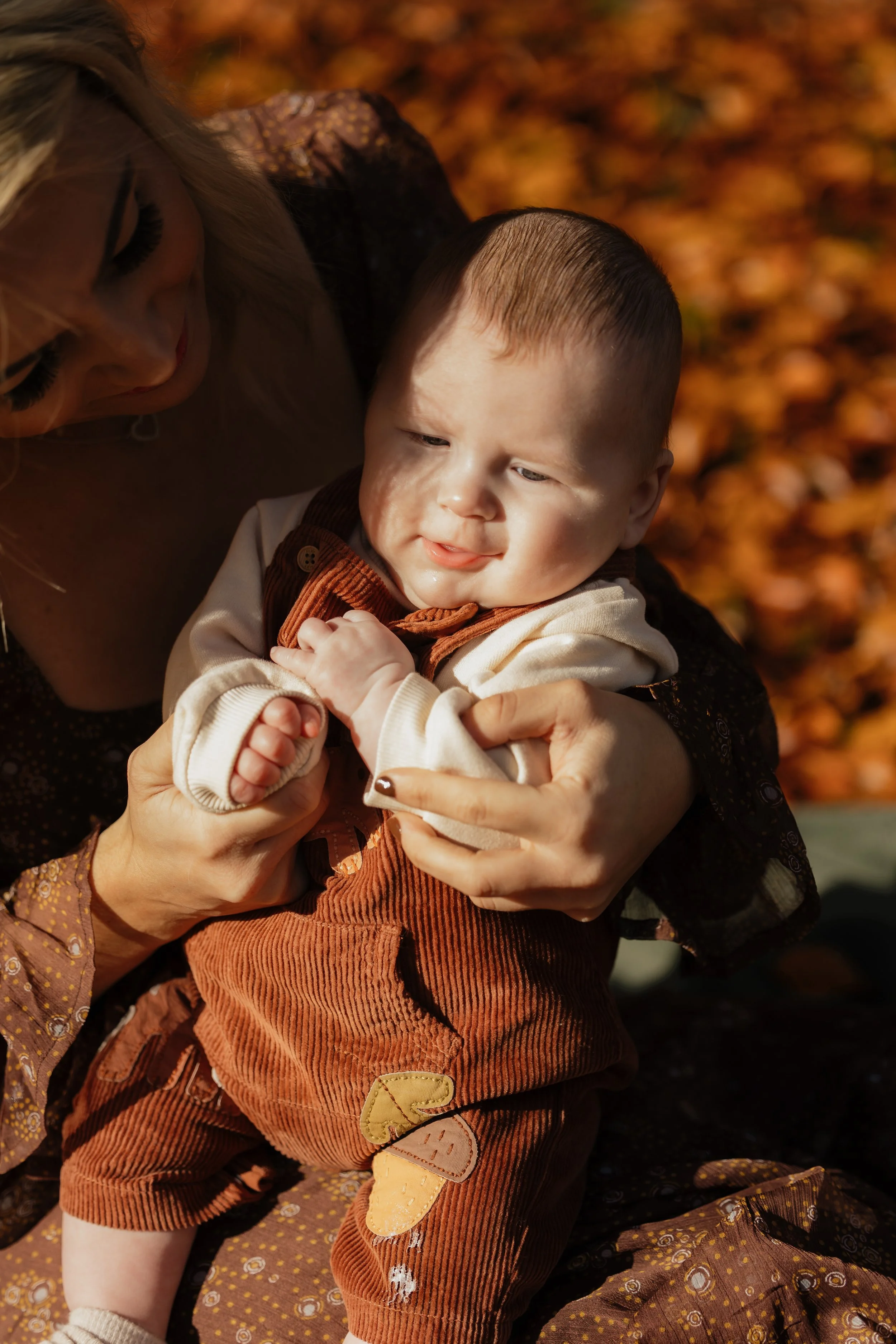 A woman holding a young boy outdoors with fall foliage in the background.