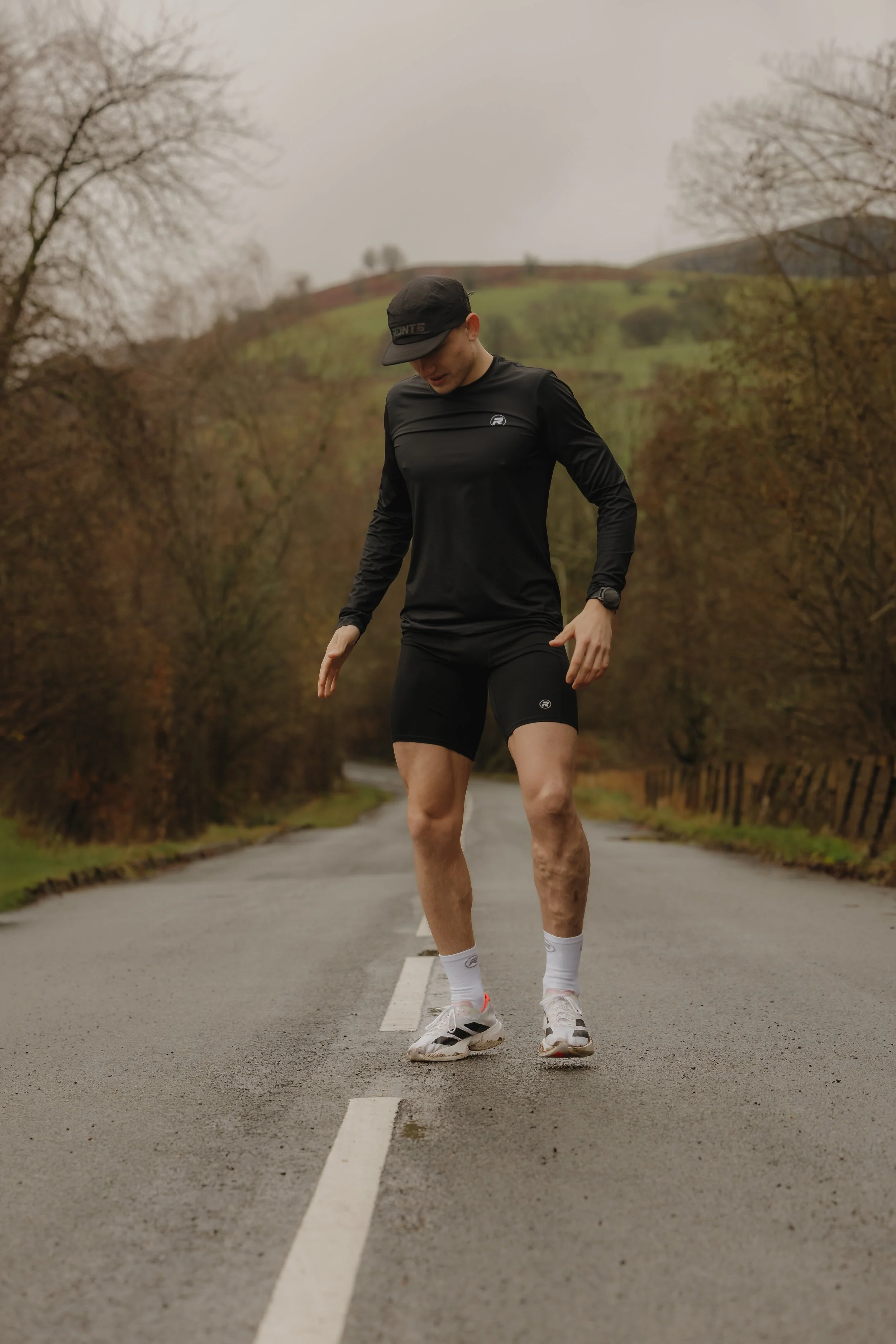 A person in athletic clothing standing on a wet road in a rural, hilly landscape with bare trees and overcast sky.