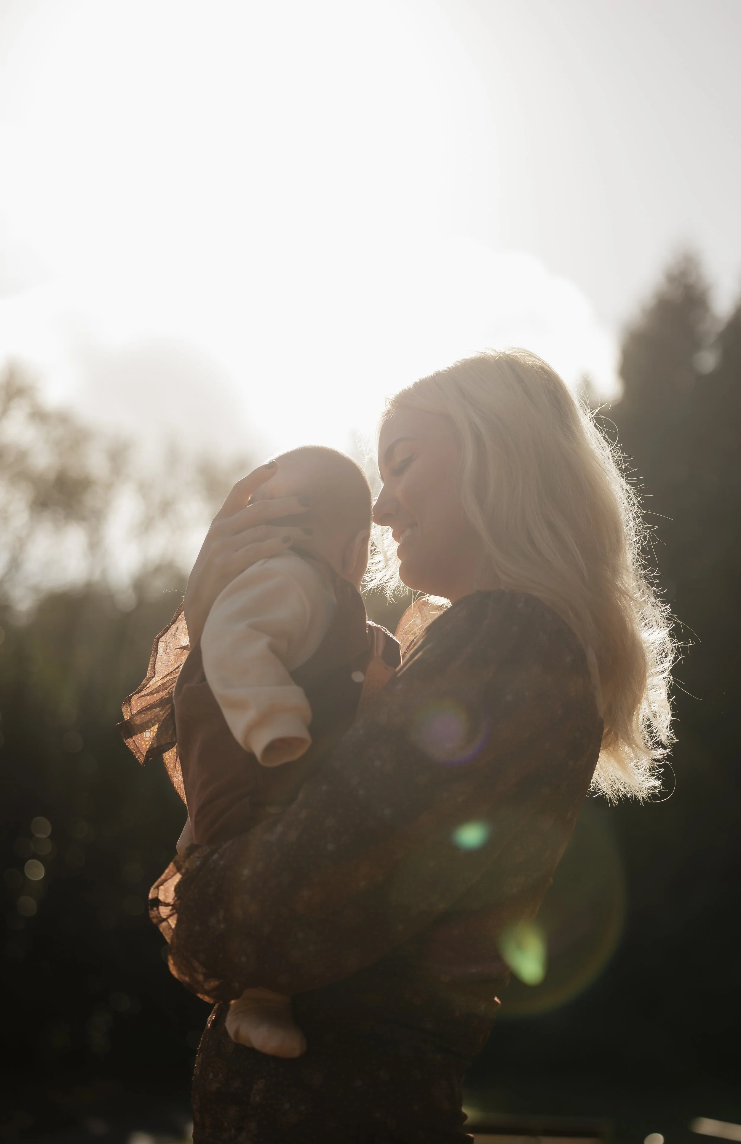 A woman holding a baby outdoors during sunset, with sunlight creating a backlit glow and lens flare.