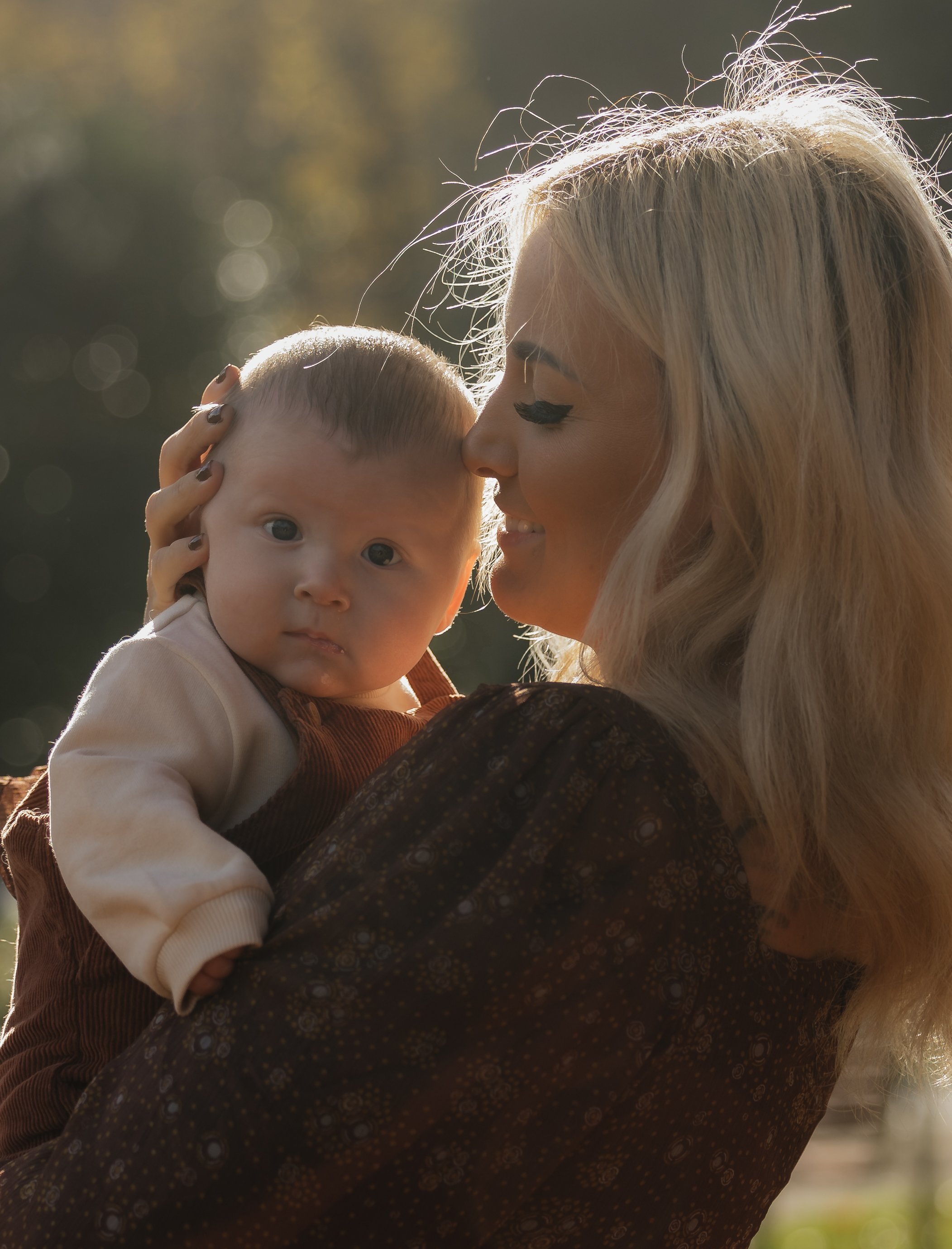 A woman holding a baby close to her face outdoors during sunset, with a blurred background of trees.