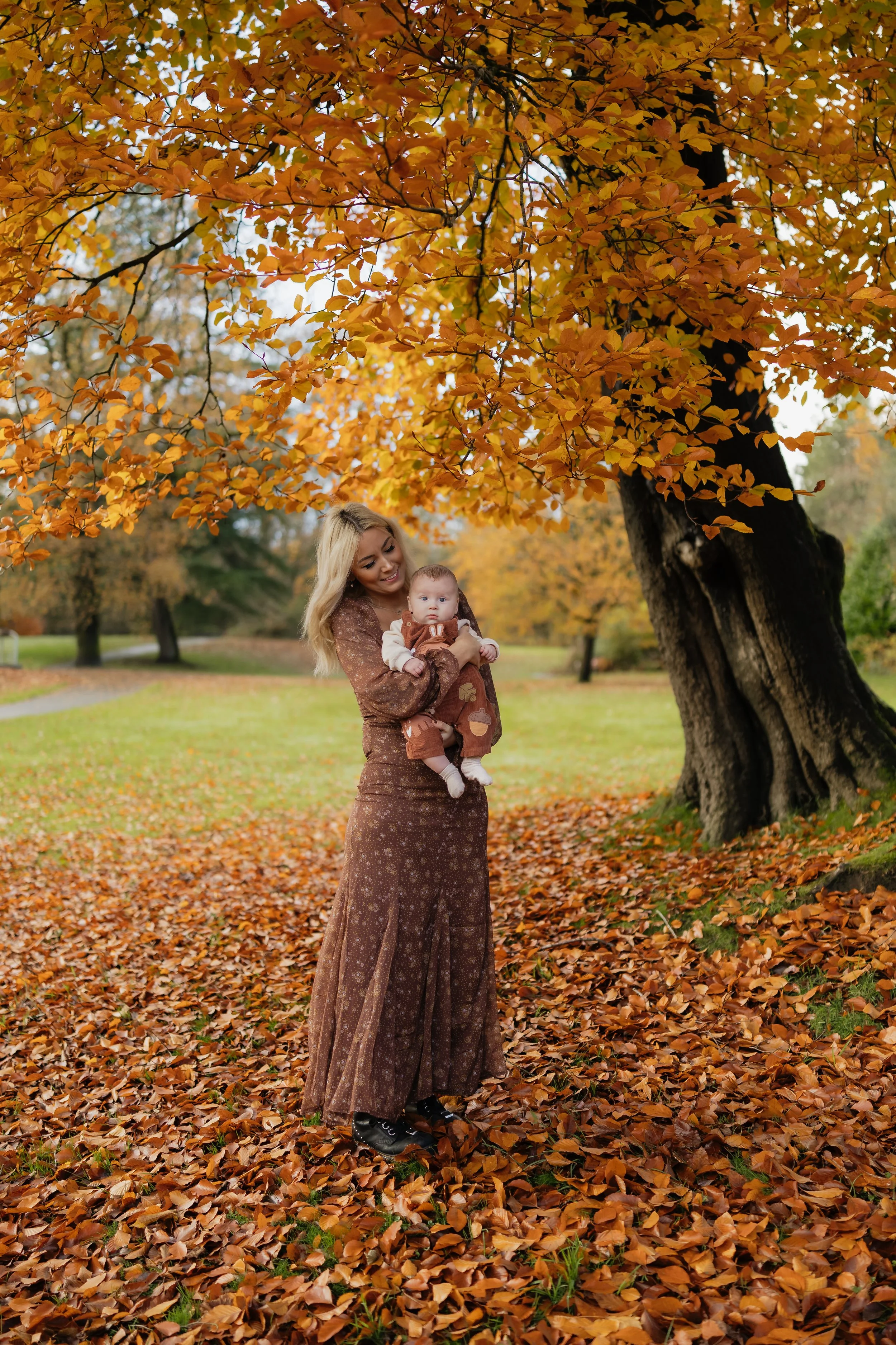 A woman holding a baby outdoors in an autumn park with a large tree and fallen leaves.