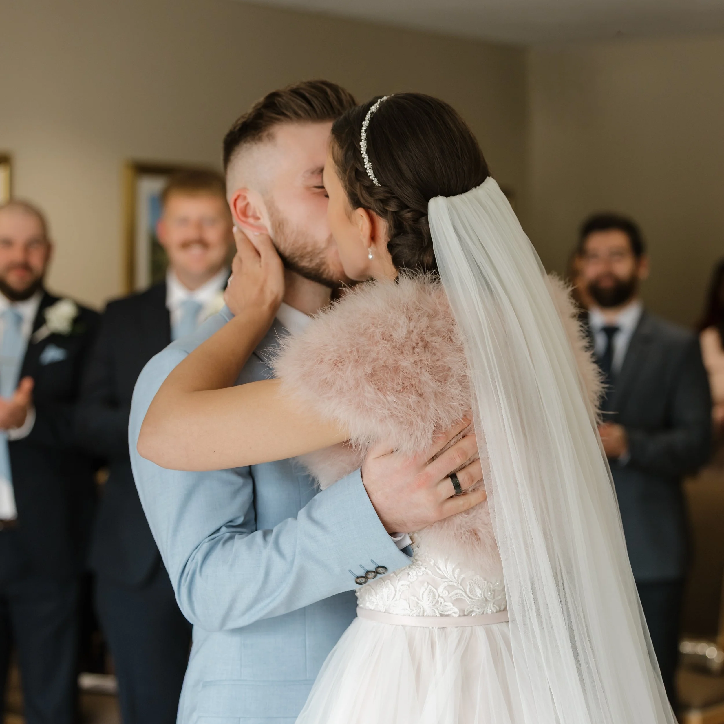 A bride and groom kiss during their wedding ceremony, with friends in the background clapping and smiling.