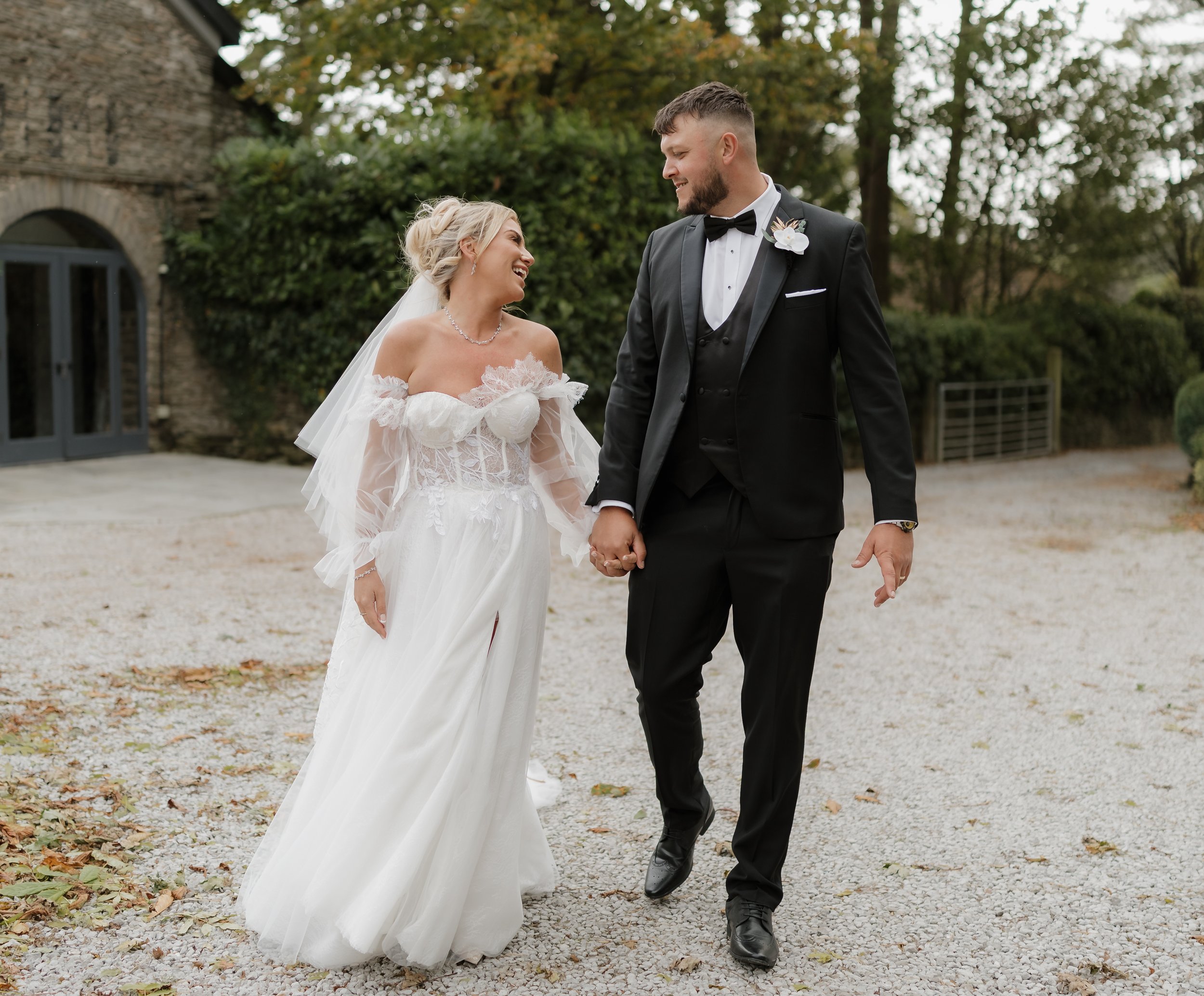 Bride and groom holding hands and smiling during their outdoor wedding ceremony, with trees and a stone building in the background.